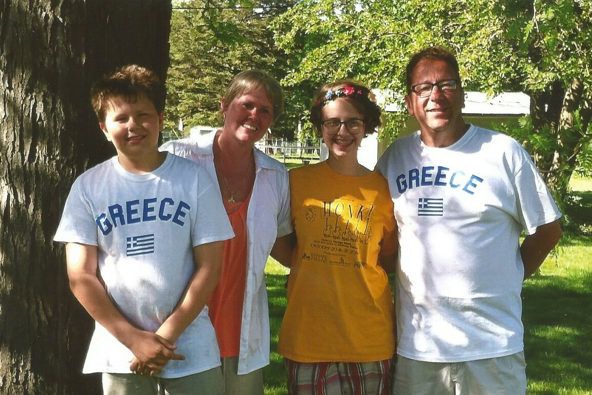 Four people posing outdoors in a park, two adults flanking two kids, all smiling on a sunny day (two ends wear Greece shirts).