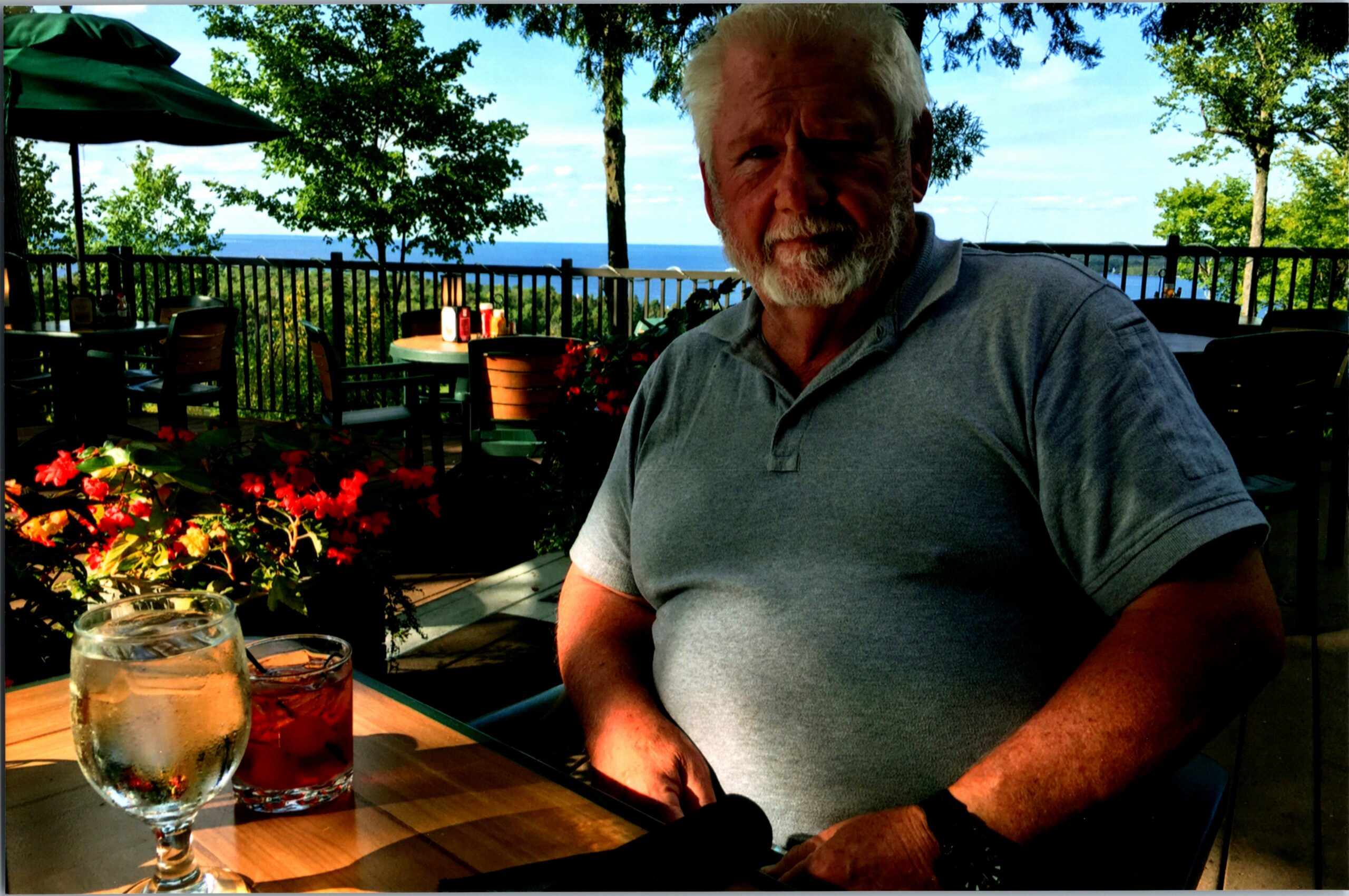 Older man with gray hair and beard sits at an outdoor cafe by the sea; drink glasses on the table in front.