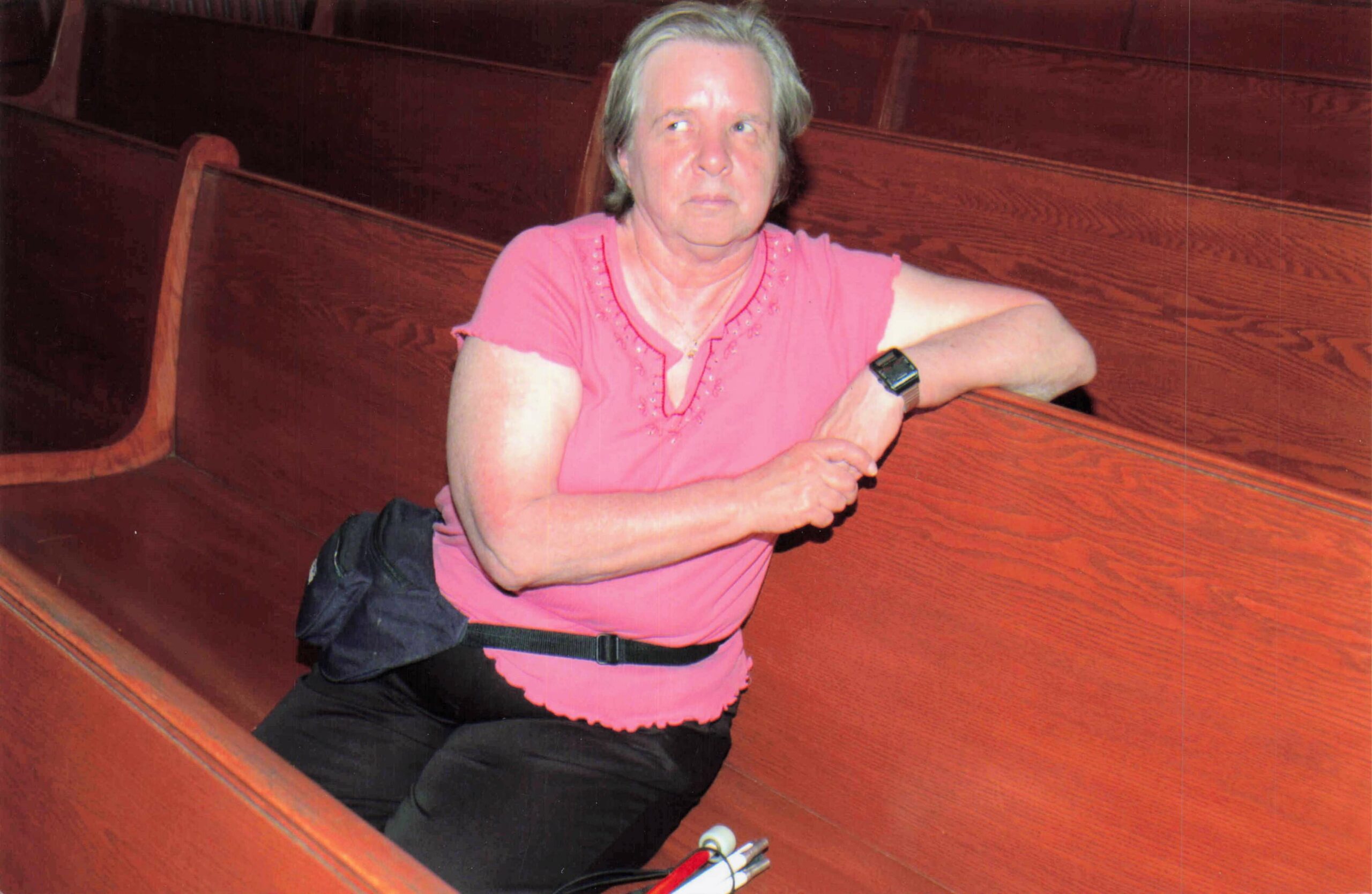 Older woman in a pink top sits on a wooden bench, resting her arms on the railing in front of her.