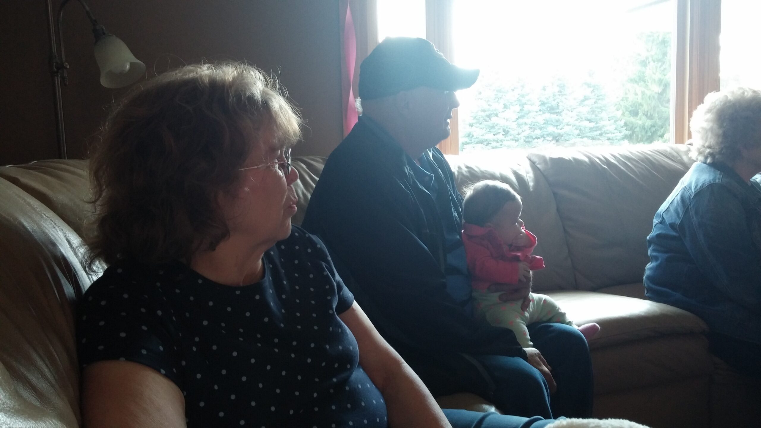 Woman with glasses in a navy polka-dot shirt sitting on a couch, looking to the right in a dim living room with a man wearing a cap and a child in a pink jacket beside him by a bright window.