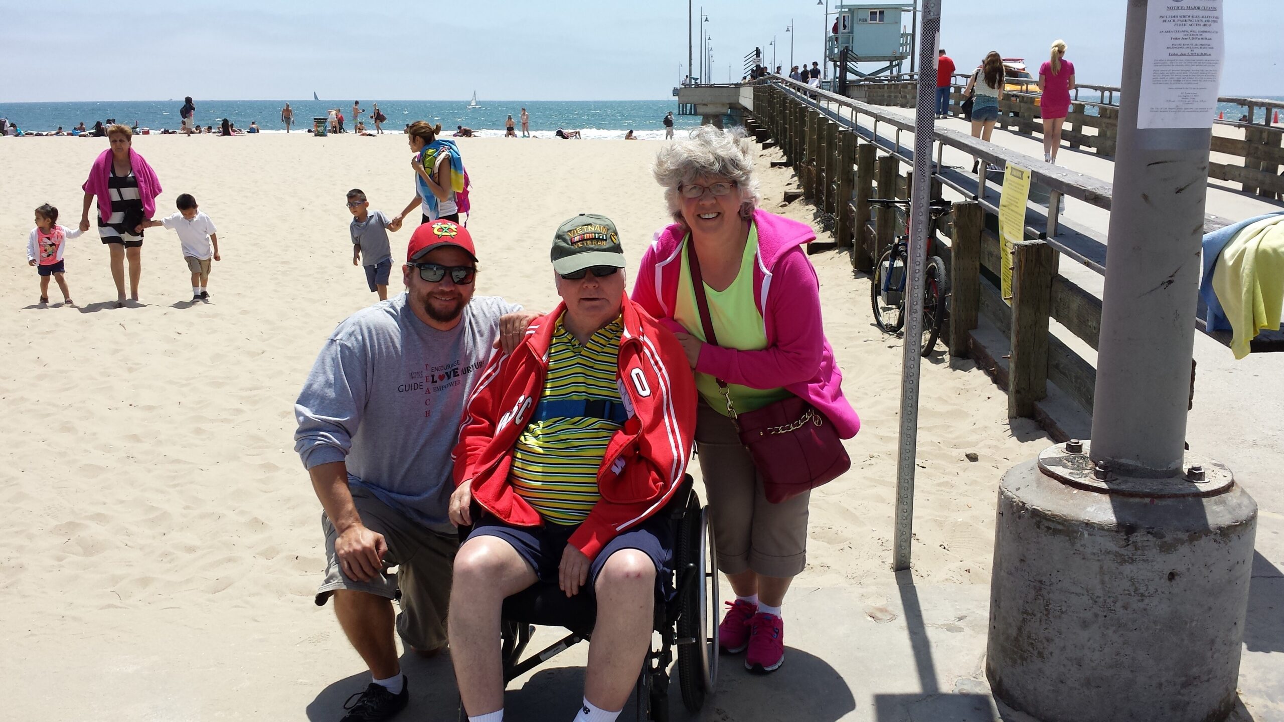 Three people at the beach posing near a wheelchair: a man in a red jacket and striped polo in a wheelchair, a kneeling man beside him, and a smiling woman in a bright pink jacket, with a boardwalk and ocean behind them.
