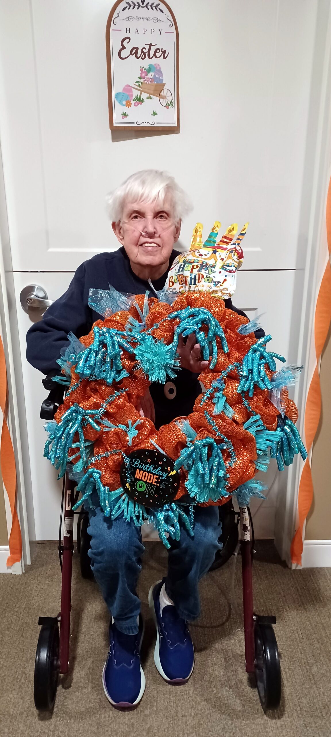 Wheelchair user with white hair holding a large orange and turquoise ribbon wreath, festive birthday badge visible, indoors with a 'Happy Easter' sign on the wall behind them.