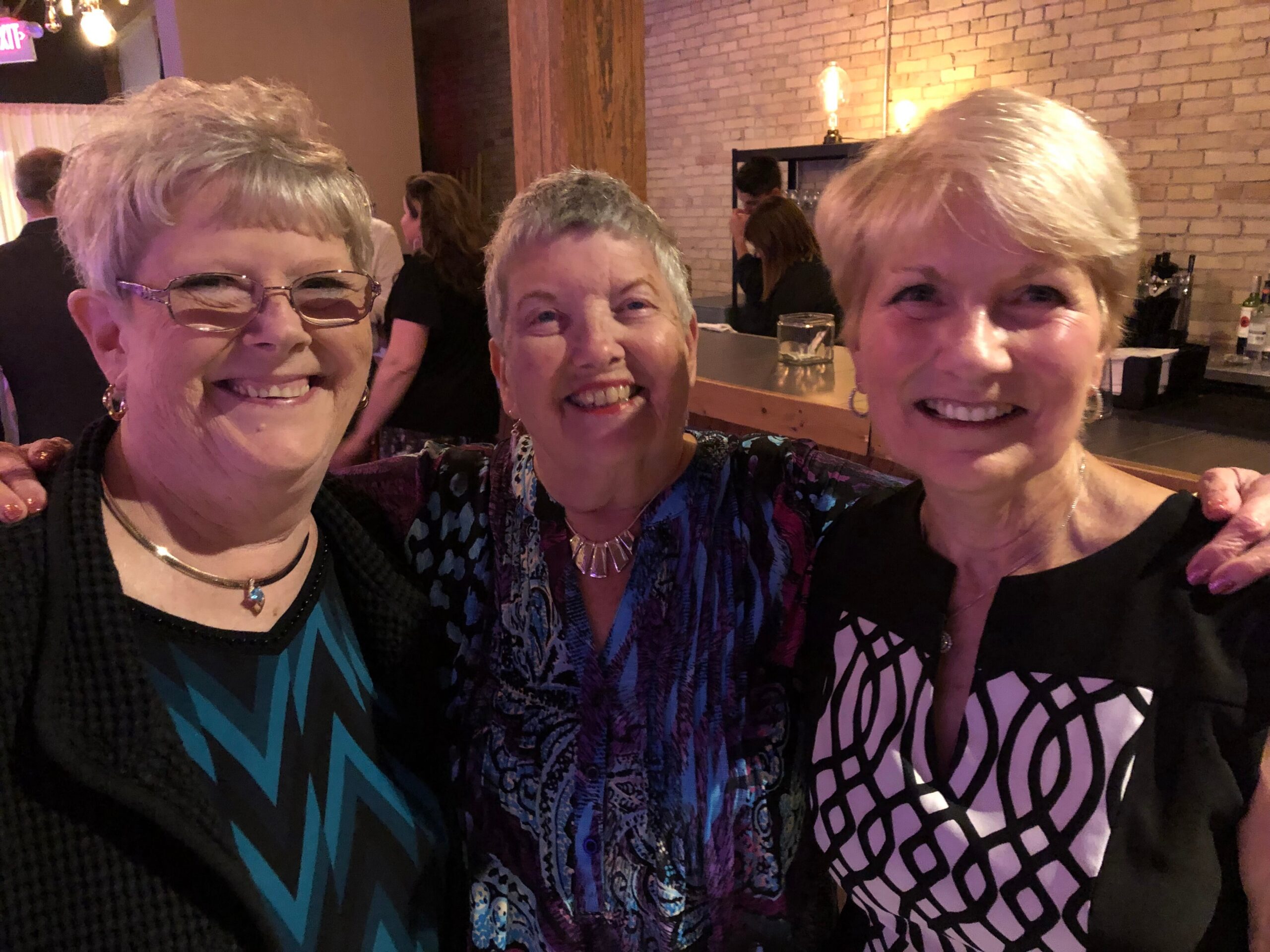 Three friends pose for a cheerful selfie at a dimly lit bar, smiling at the camera with arms around each other.