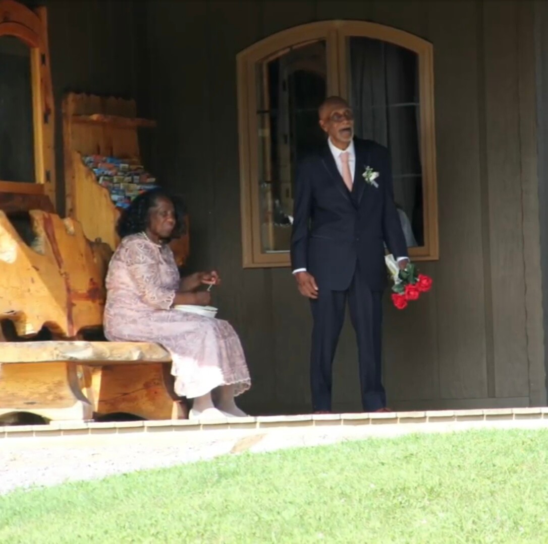 A man in a dark suit with a pink tie stands on a wooden porch holding a bouquet of red roses, while a woman in a light pink lace dress sits on a bench nearby.