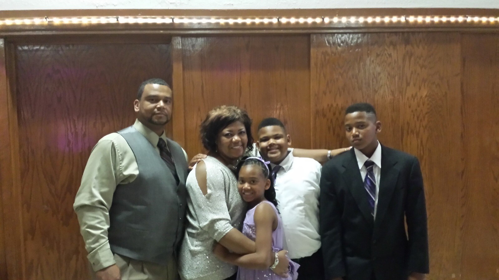 Family of five posing together in formal attire in front of a wood-paneled wall at a event place.