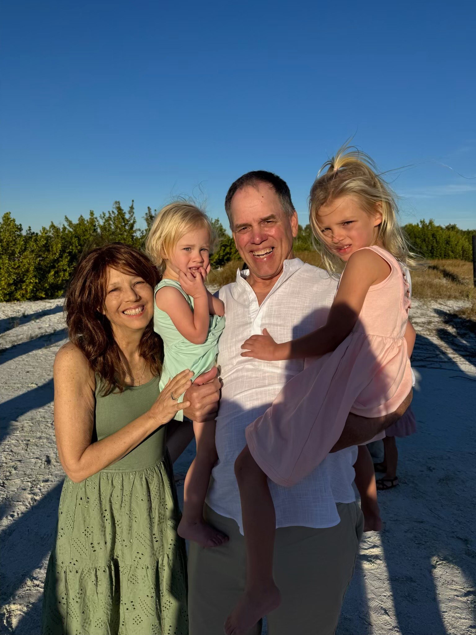 A smiling family of four posing outdoors on a sunny day, with a clear blue sky and sandy ground behind them.