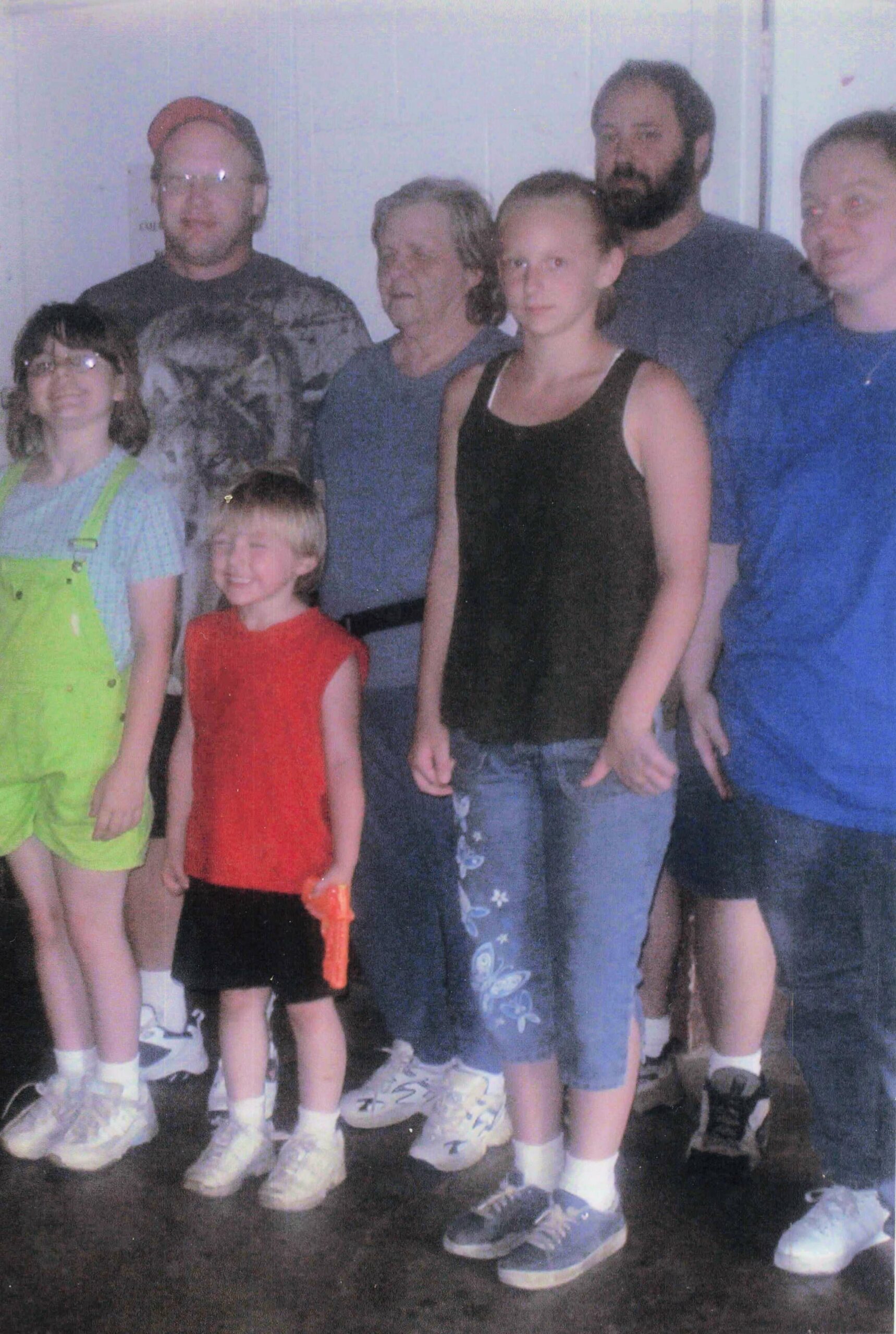 Group of seven people posing indoors for a casual photo, with two children in front and adults behind them.