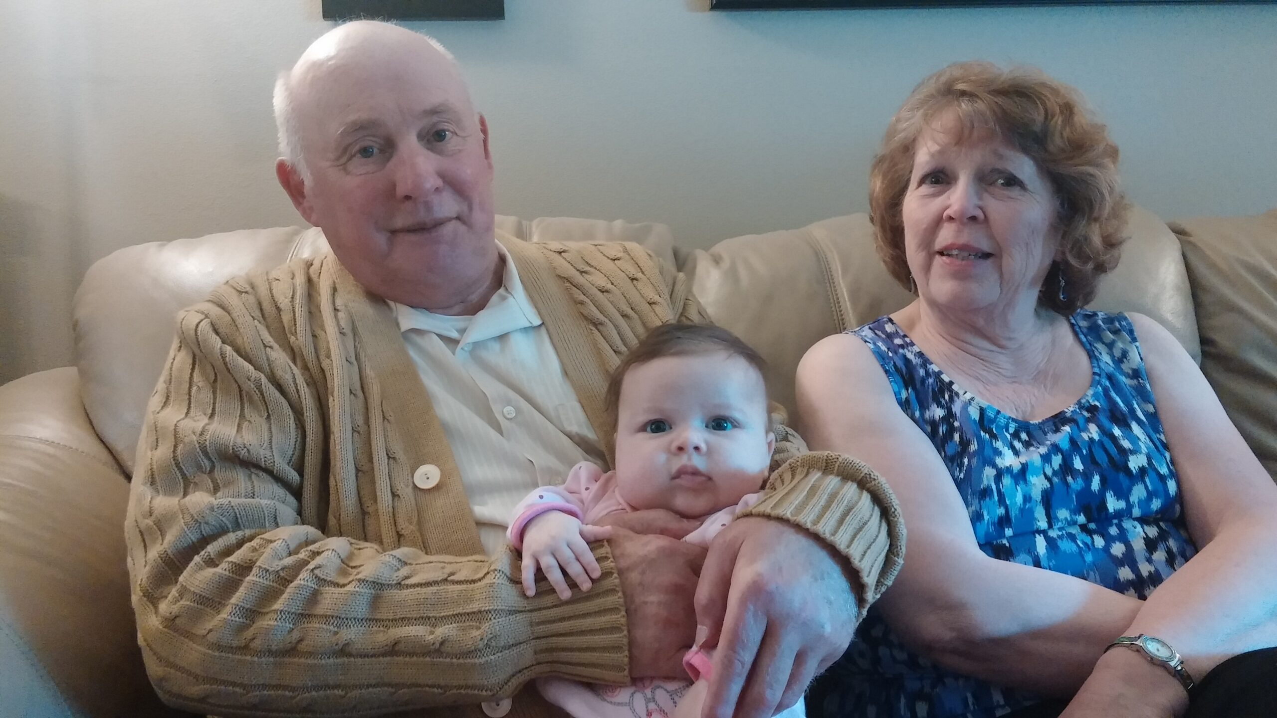 Smiling older man and woman sit on a beige couch with a baby in the man's arms on their laps.