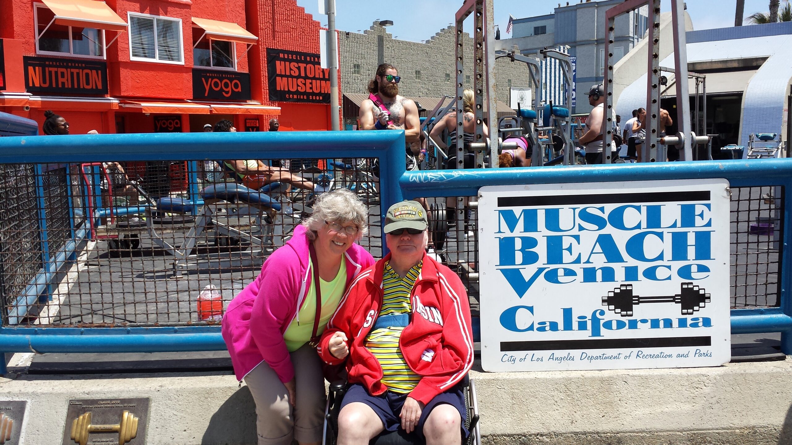 Two smiling people in bright jackets pose at Muscle Beach Venice, with a blue railing and sign reading 'Muscle Beach Venice California' behind them, sunny boardwalk scene.