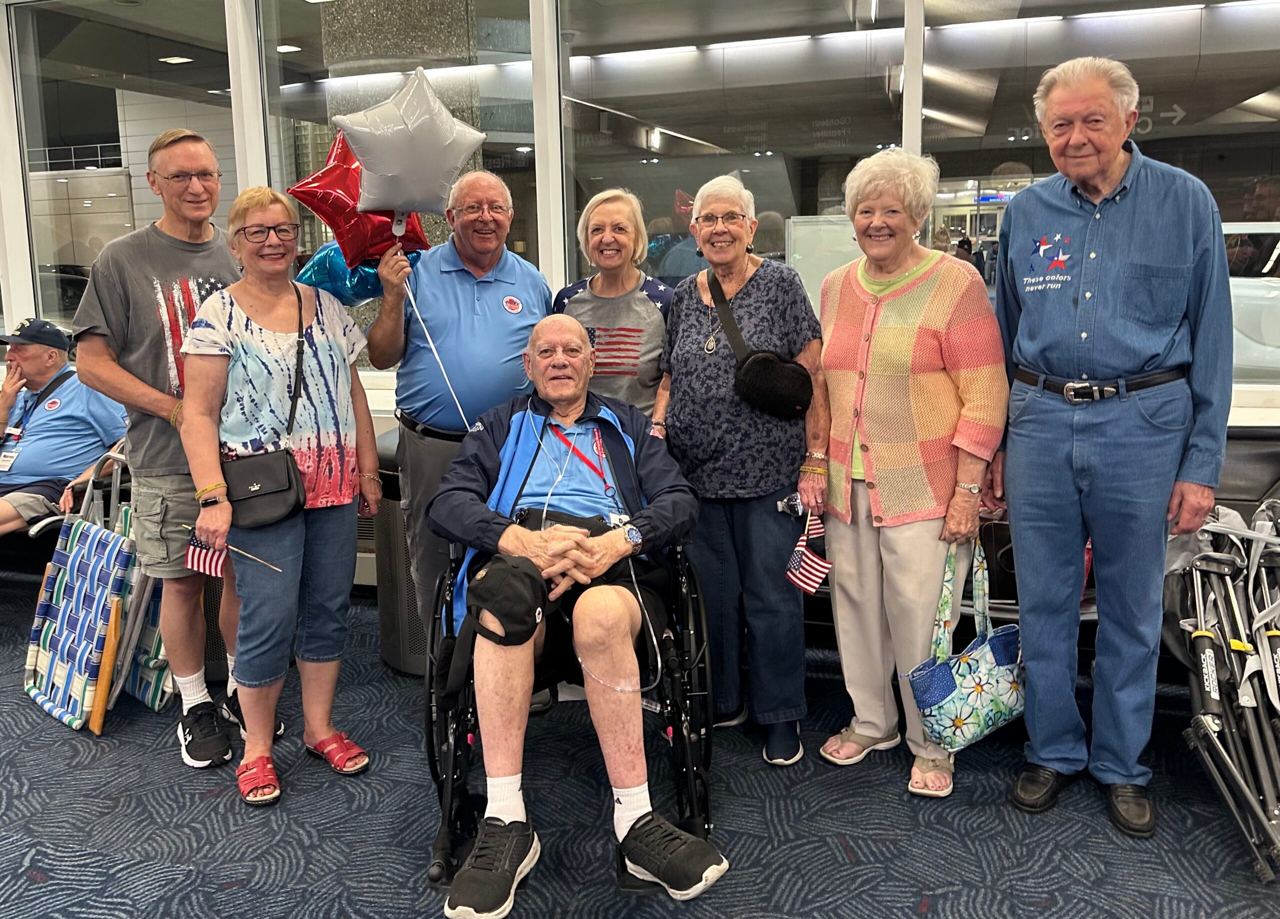 Eight seniors posing for a photo in an airport or terminal, some holding red, white, and blue balloons and American flags. creased patriotic attire and luggage visible in the background.