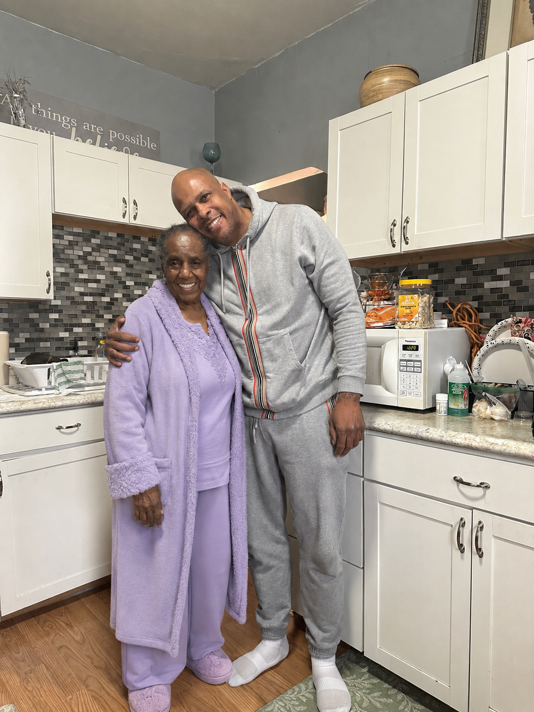 Smiling elderly woman in a lavender robe stands with a man in a gray tracksuit in a bright kitchen, arms around each other.