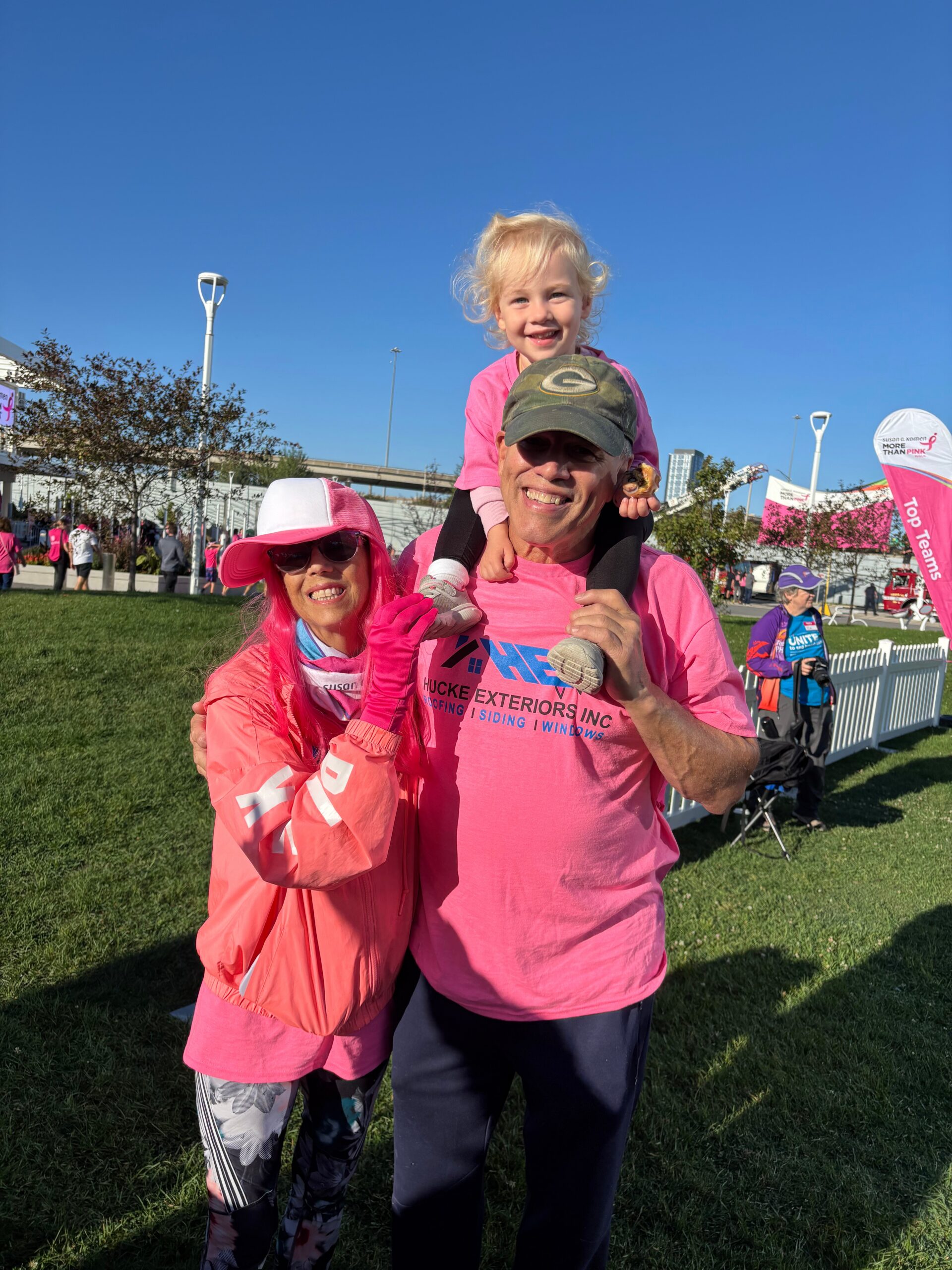 Man in a pink shirt carries a smiling child on his shoulders at an outdoor charity event; people and tents in the background.