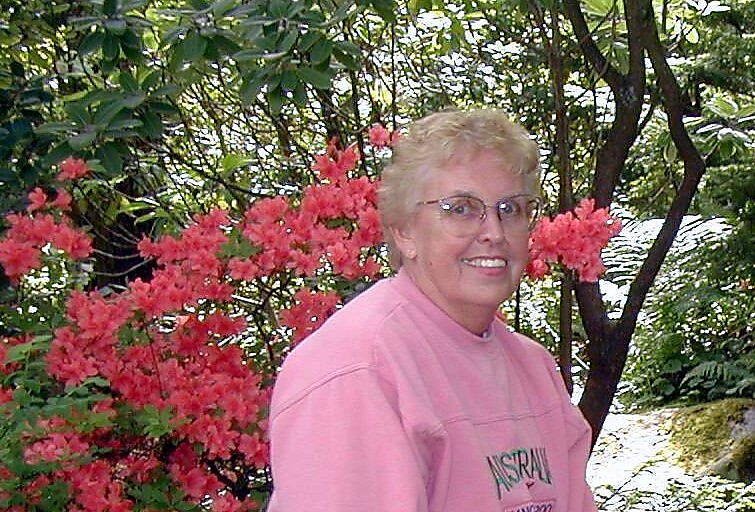 Older woman with short blond hair wearing a pink shirt, smiling, in front of pink azalea bushes in a garden.