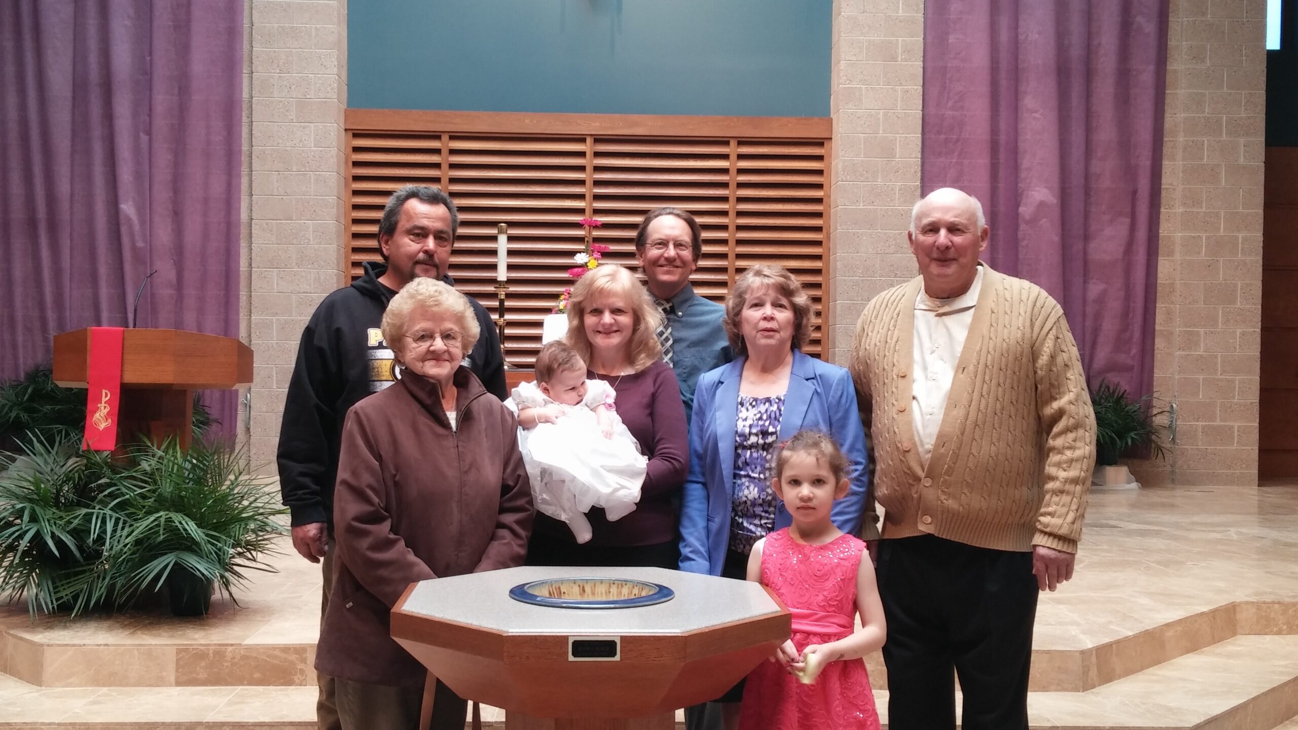 Group of seven people posing around a baptismal font in a church, with a baby dressed in white at the center.