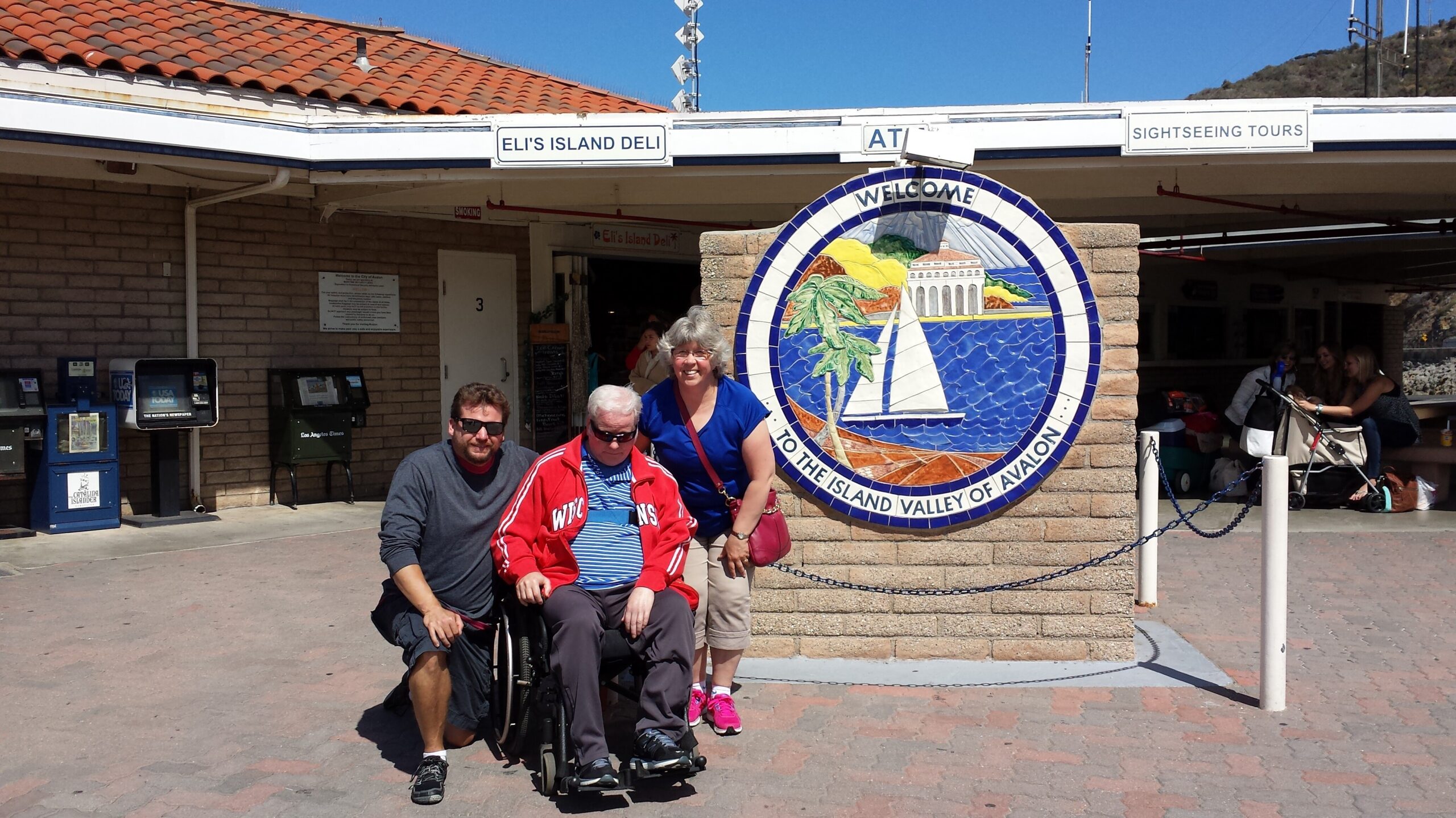 Three people pose in front of a large circular mosaic sign that reads 'Welcome to the Island Valley of Avalon,' featuring a sailboat and palm tree, beside Eli's Island Deli.