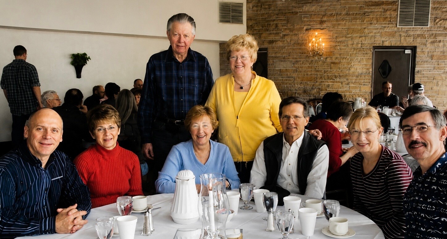 Eight adults sit and stand around a round dining table, smiling for the photo in a restaurant setting with a white tablecloth and glassware nearby.