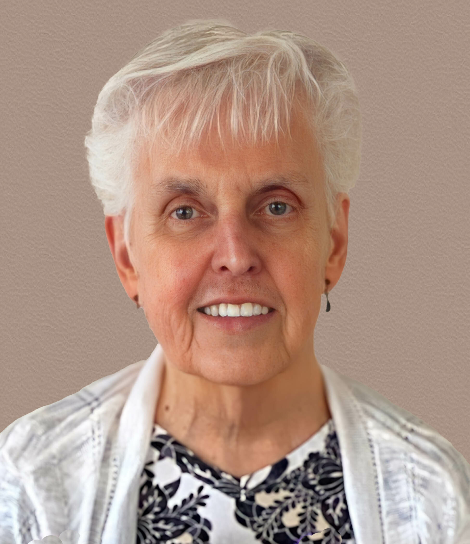 Close-up portrait of an older person with short white hair smiling, wearing a patterned black-and-white shirt and a light jacket against a neutral background.