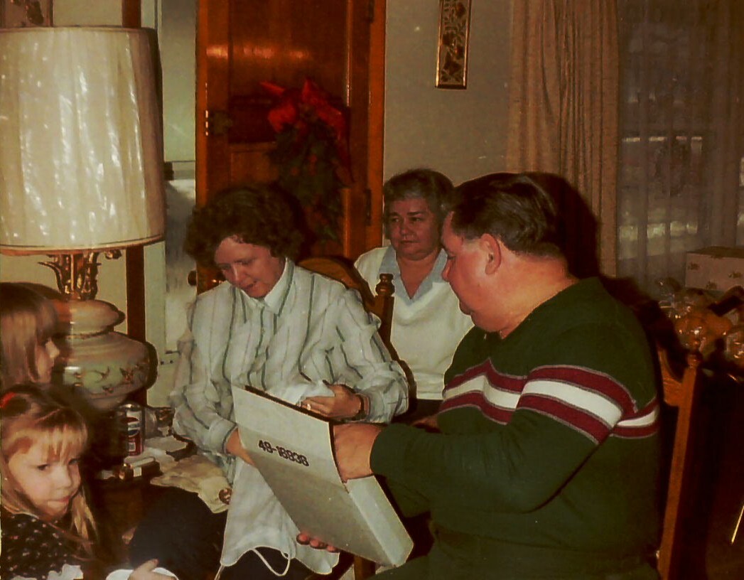 Family at a dining table; a man shows a large envelope or folder to a woman while a young girl watches nearby.