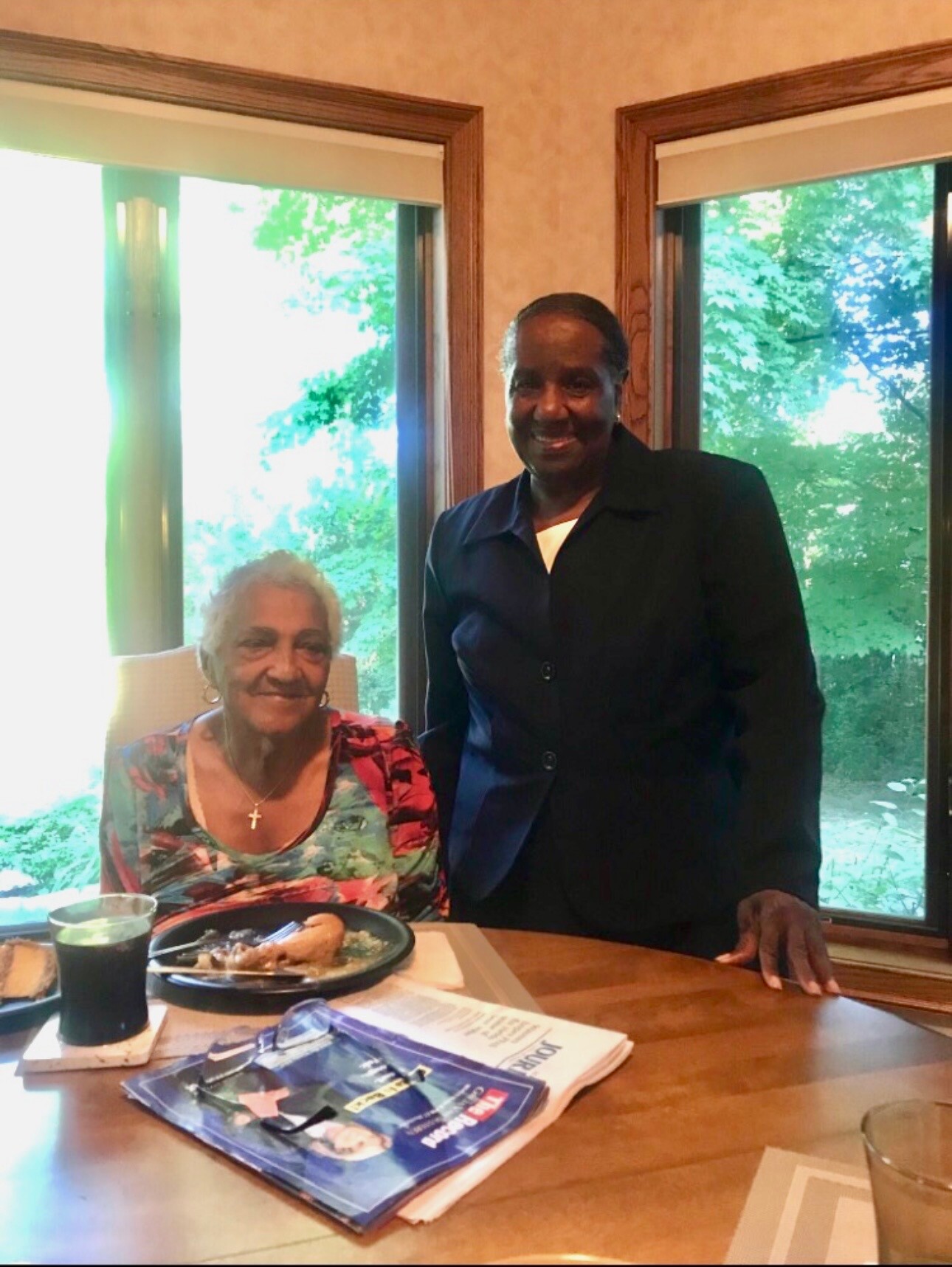Smiling elderly woman seated at a dining table with a plate of food, while a man in a dark suit stands beside her by large windows with greenery outside.