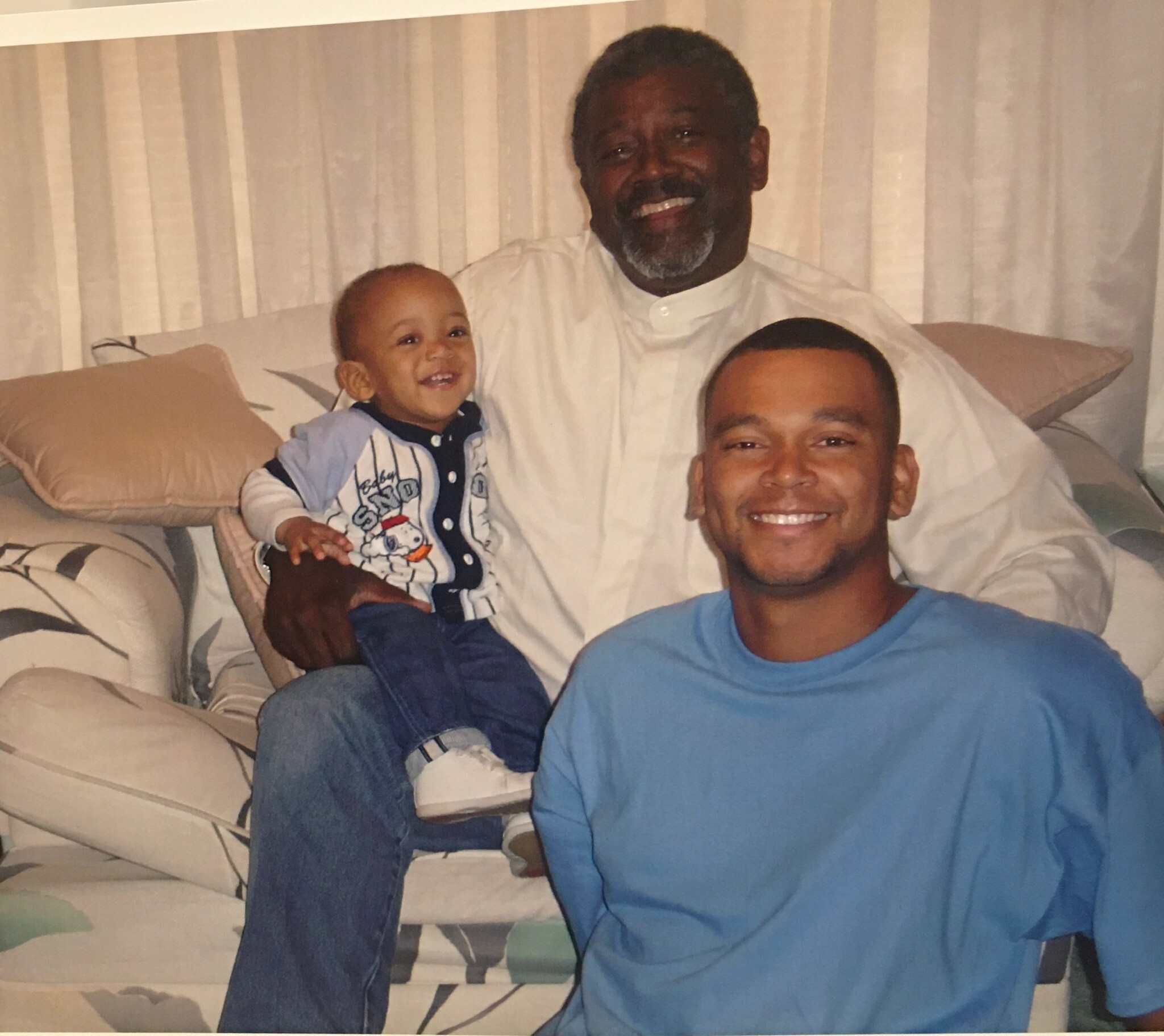 Smiling man in a blue shirt sits on a couch with a baby on his lap and another man in white beside them, all posing closely together on a sofa.