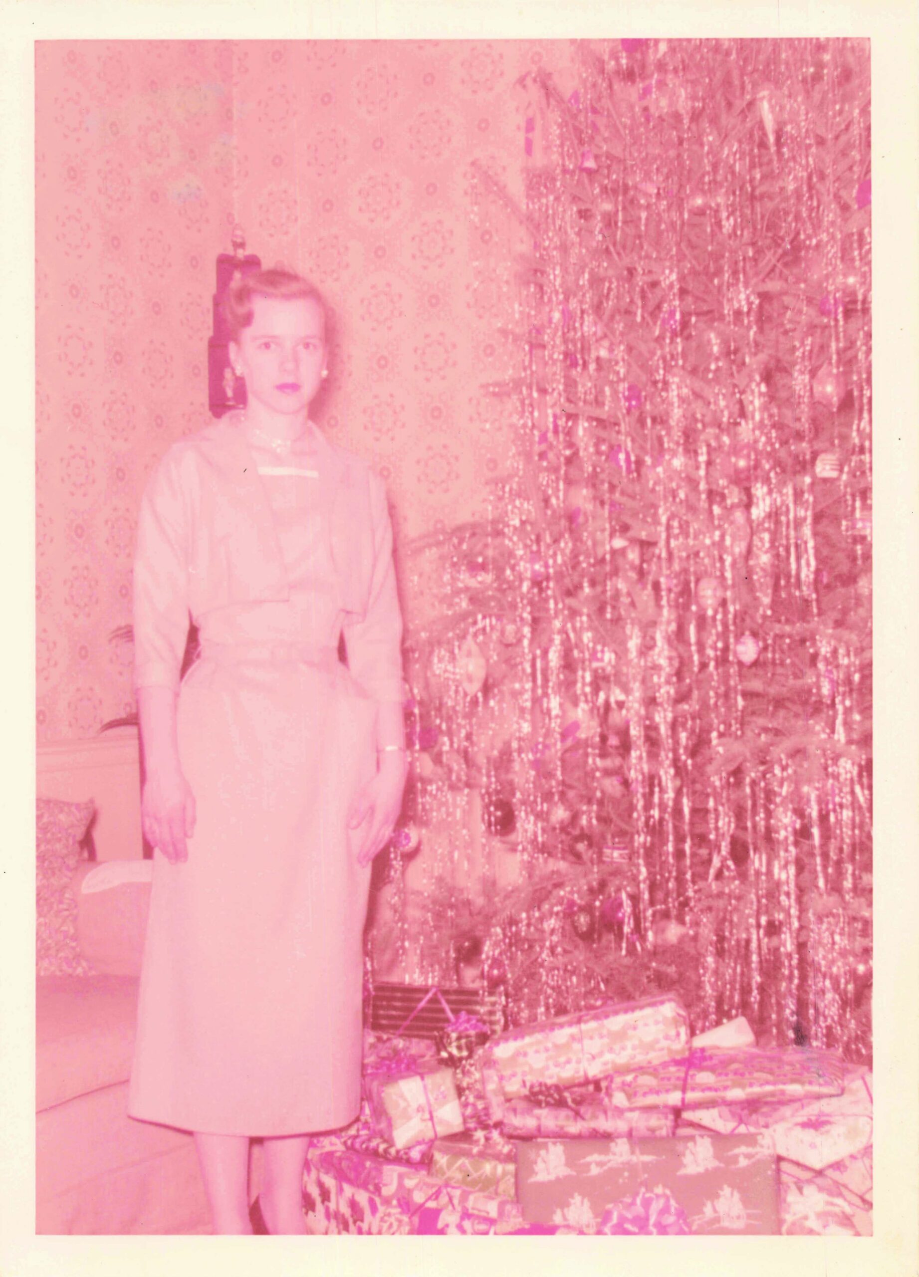 Woman in a pale dress standing indoors beside a decorated Christmas tree with wrapped presents beneath it.