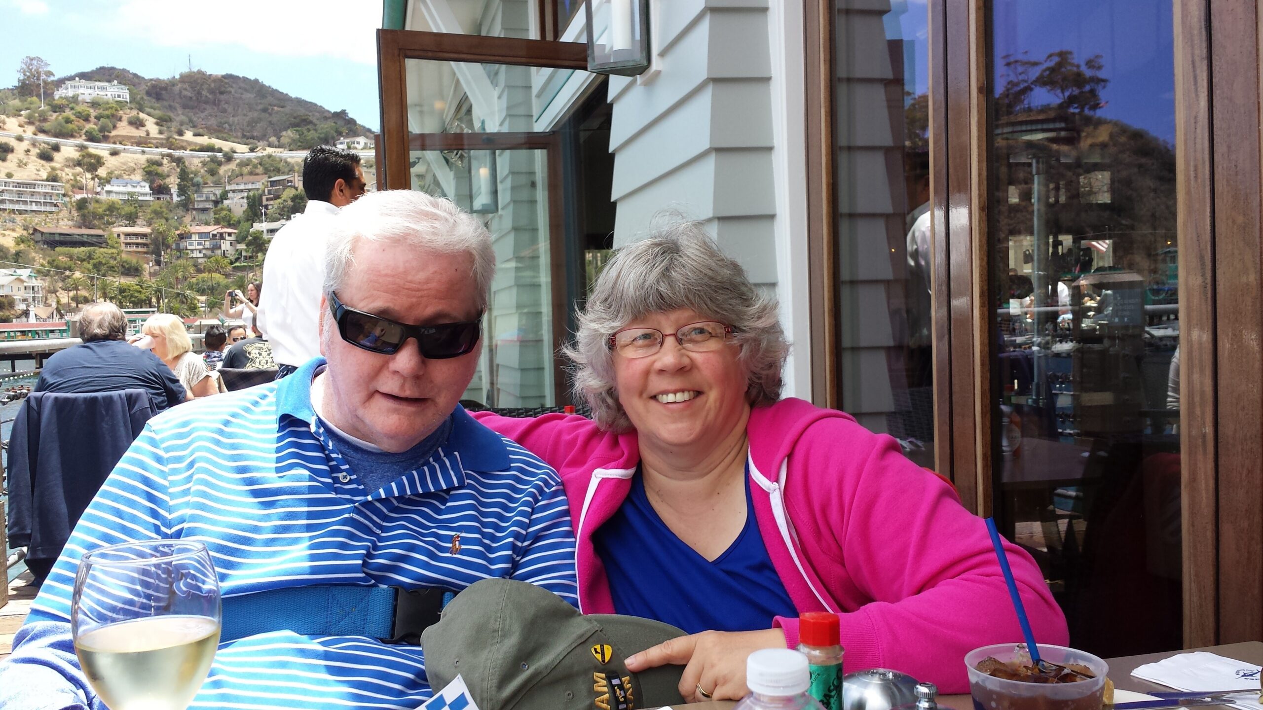 Two adults sit together at an outdoor restaurant table, smiling with drinks and food in front, hillside town in the background