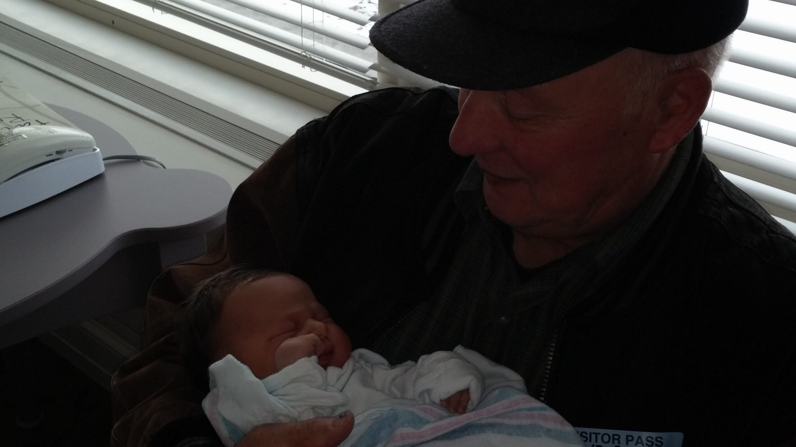 Older man wearing a cap cradling a newborn wrapped in a striped blanket by a windowed wall.