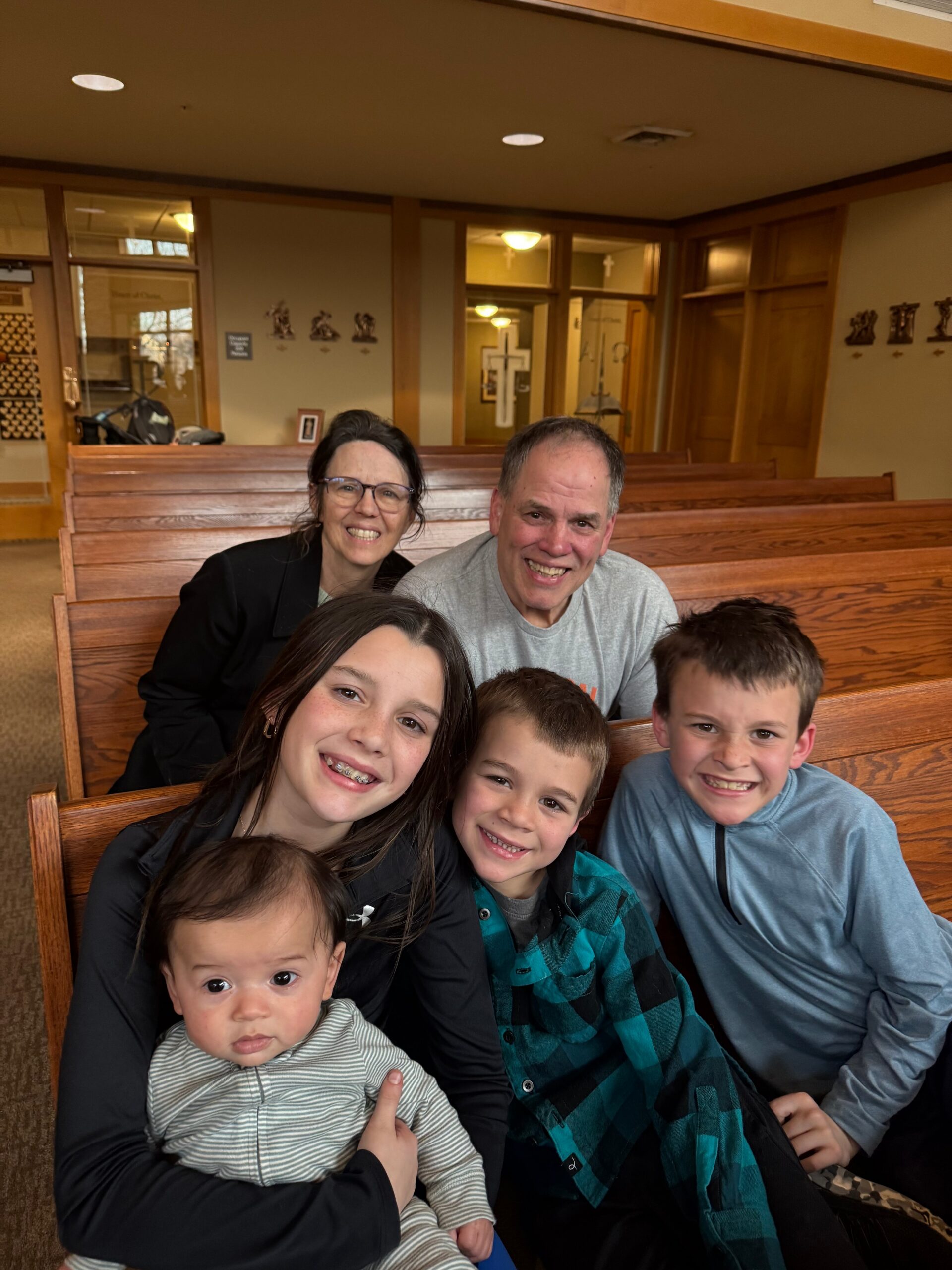 Six family members sit closely on wooden church pews, smiling at the camera for a group photo inside a church.