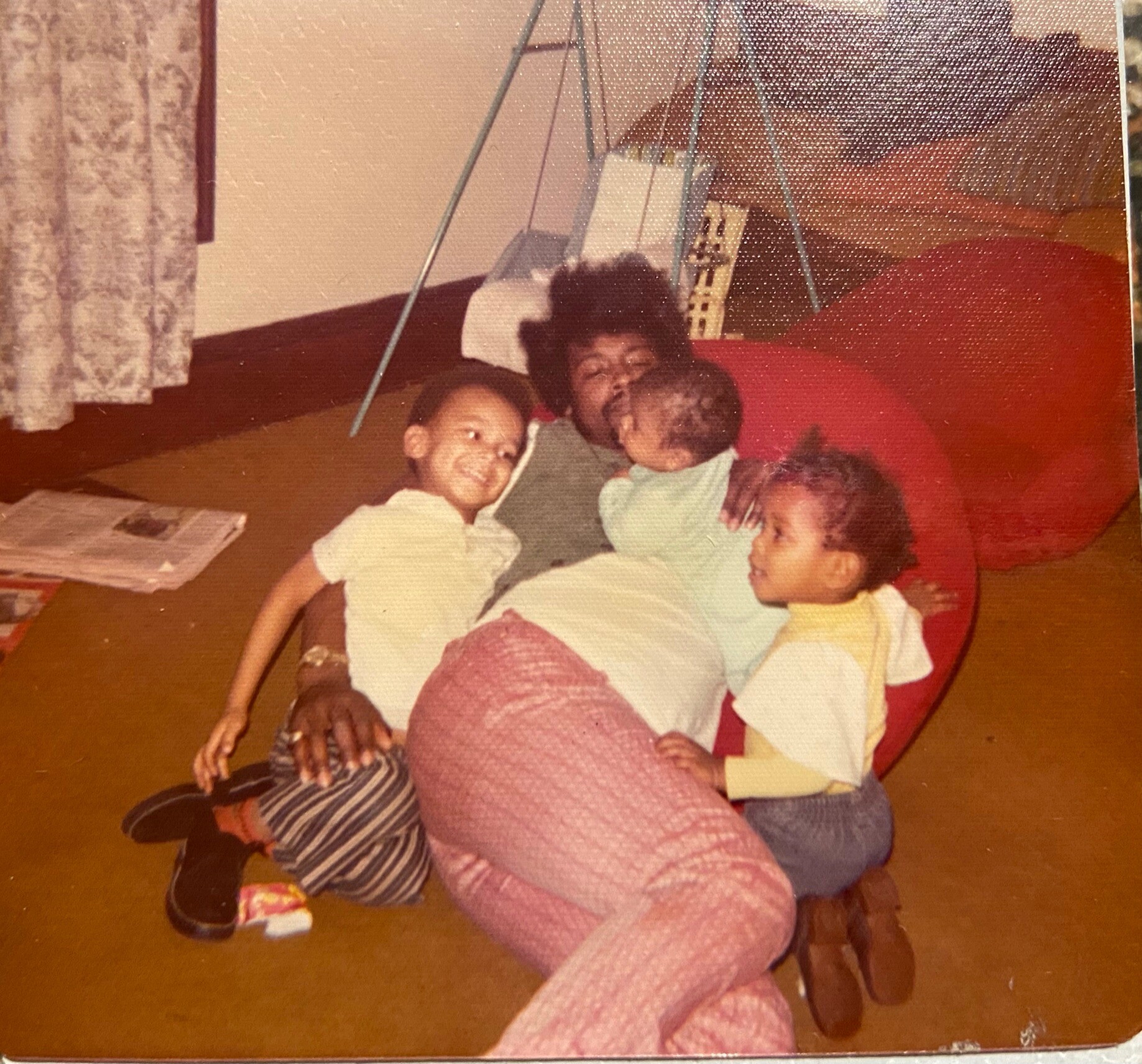Several children sit on a brown carpet indoors, hugging and smiling, with a metal-framed swing set behind them.