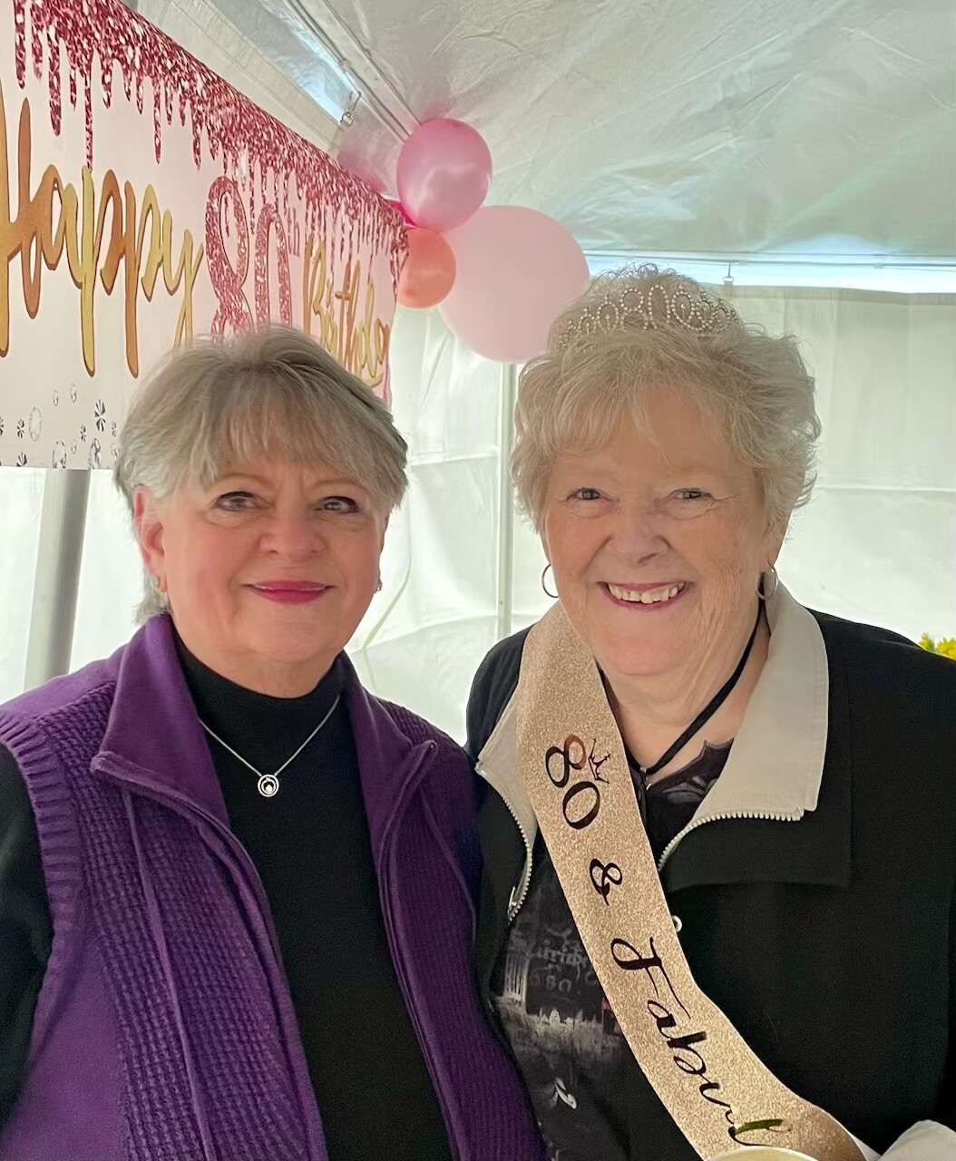 Two smiling women pose at a birthday celebration; one wears a tiara and a beige sash reading '80th Birthday'.