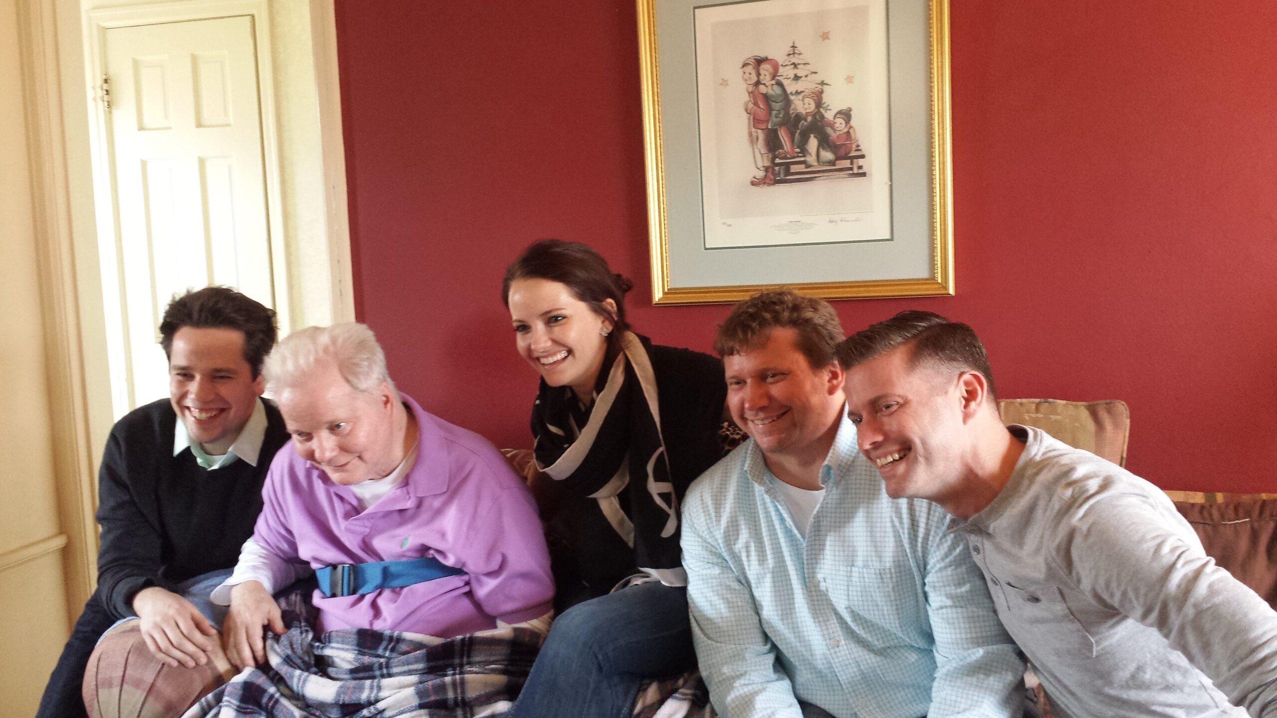 Five adults sit on a sofa in a living room, smiling for a group photo with a red wall and framed artwork behind them.