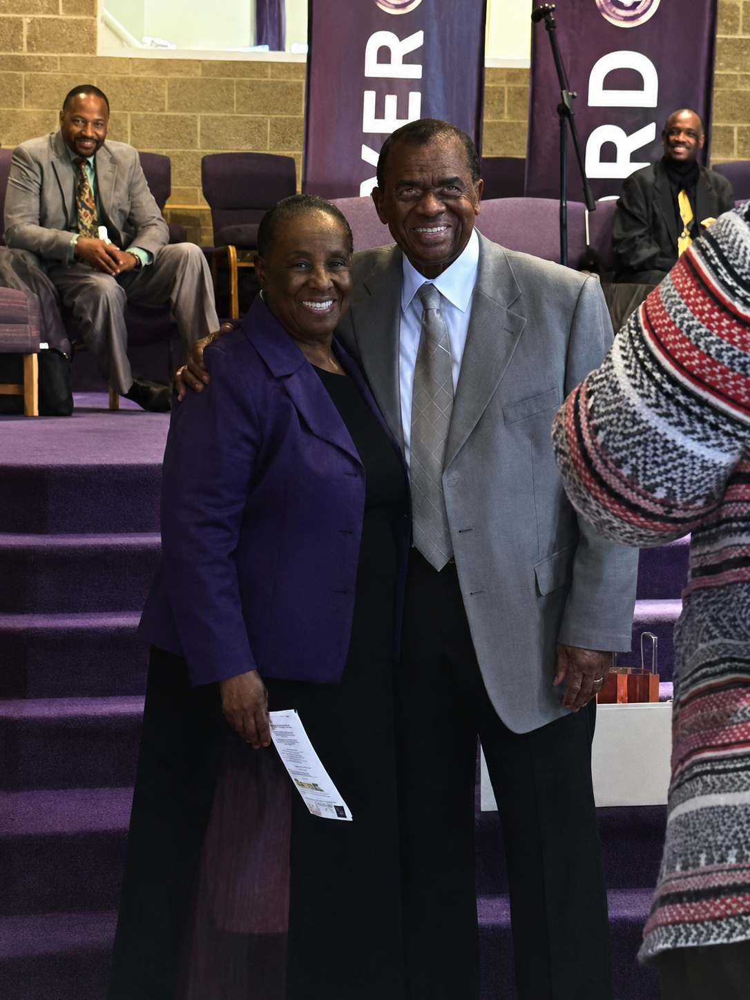 Couple posing together on a purple-carpeted stage, smiling; woman wearing a purple blazer and black dress, man in a gray suit and tie, with banners in the background to the stage left and right.