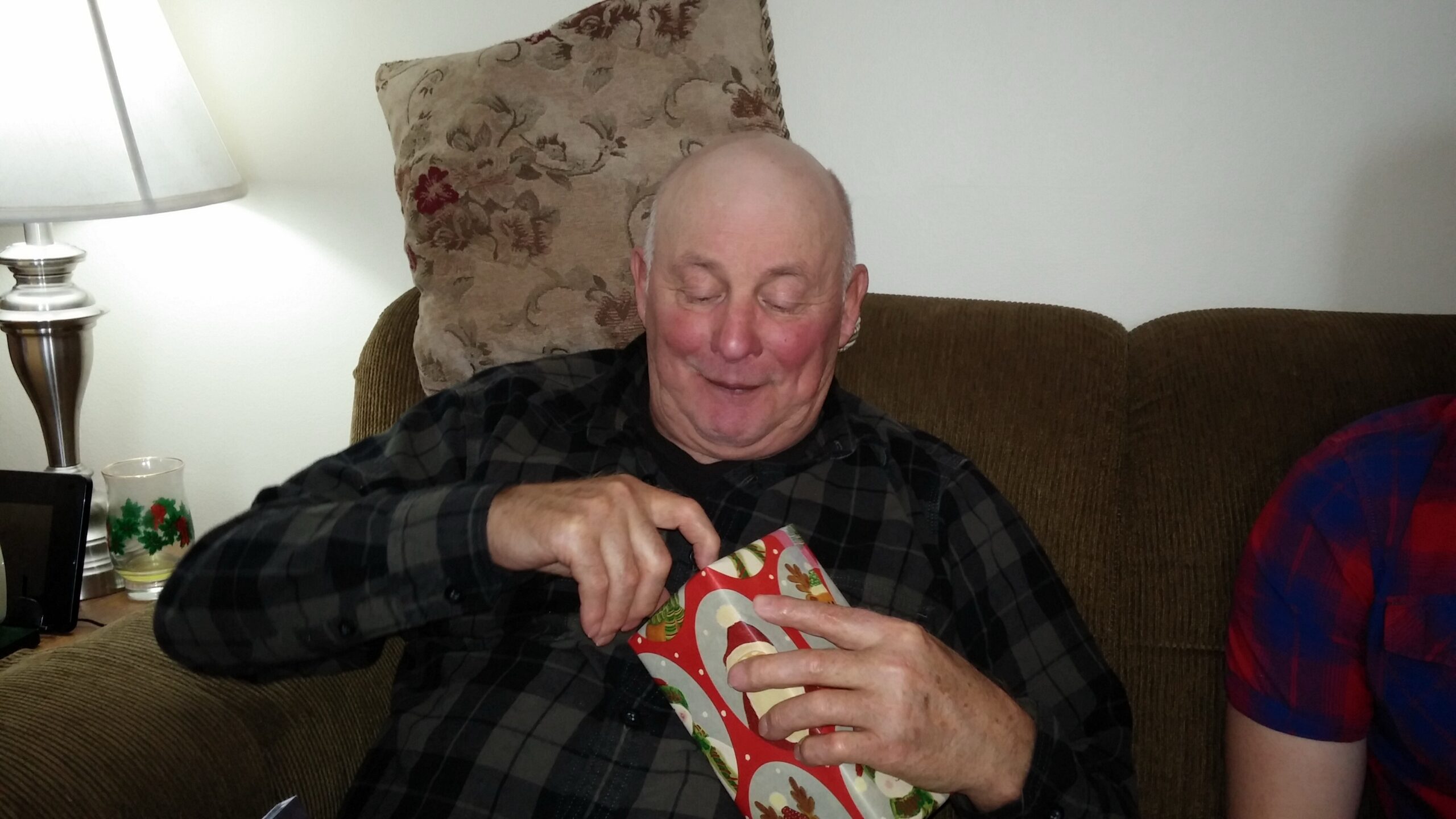 Older man with a smile tearing open a Christmas gift while sitting on a brown couch, festive wrapping paper in hand.