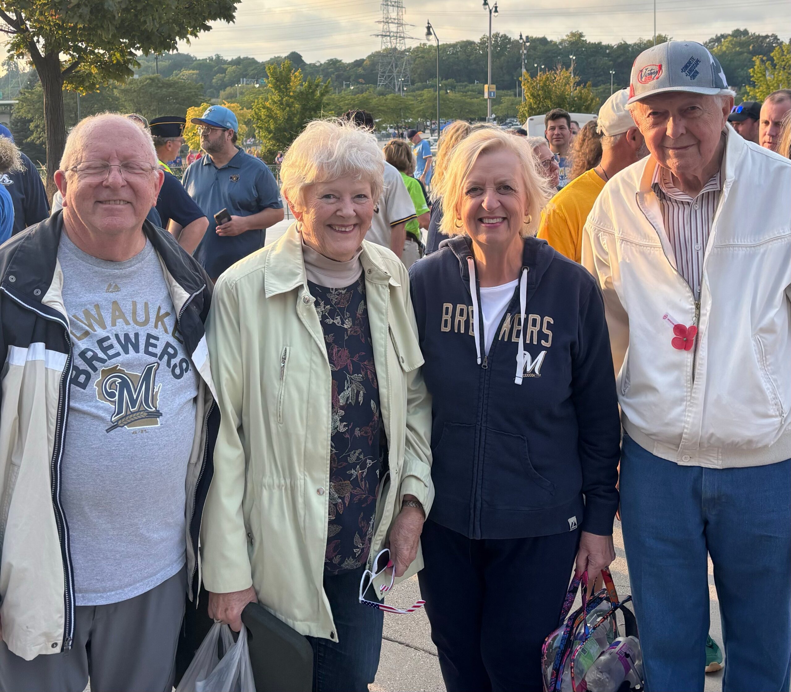 Three smiling seniors stand together at an outdoor event, each holding a bag, in daylight with trees in the background strung with event crowds nearby.