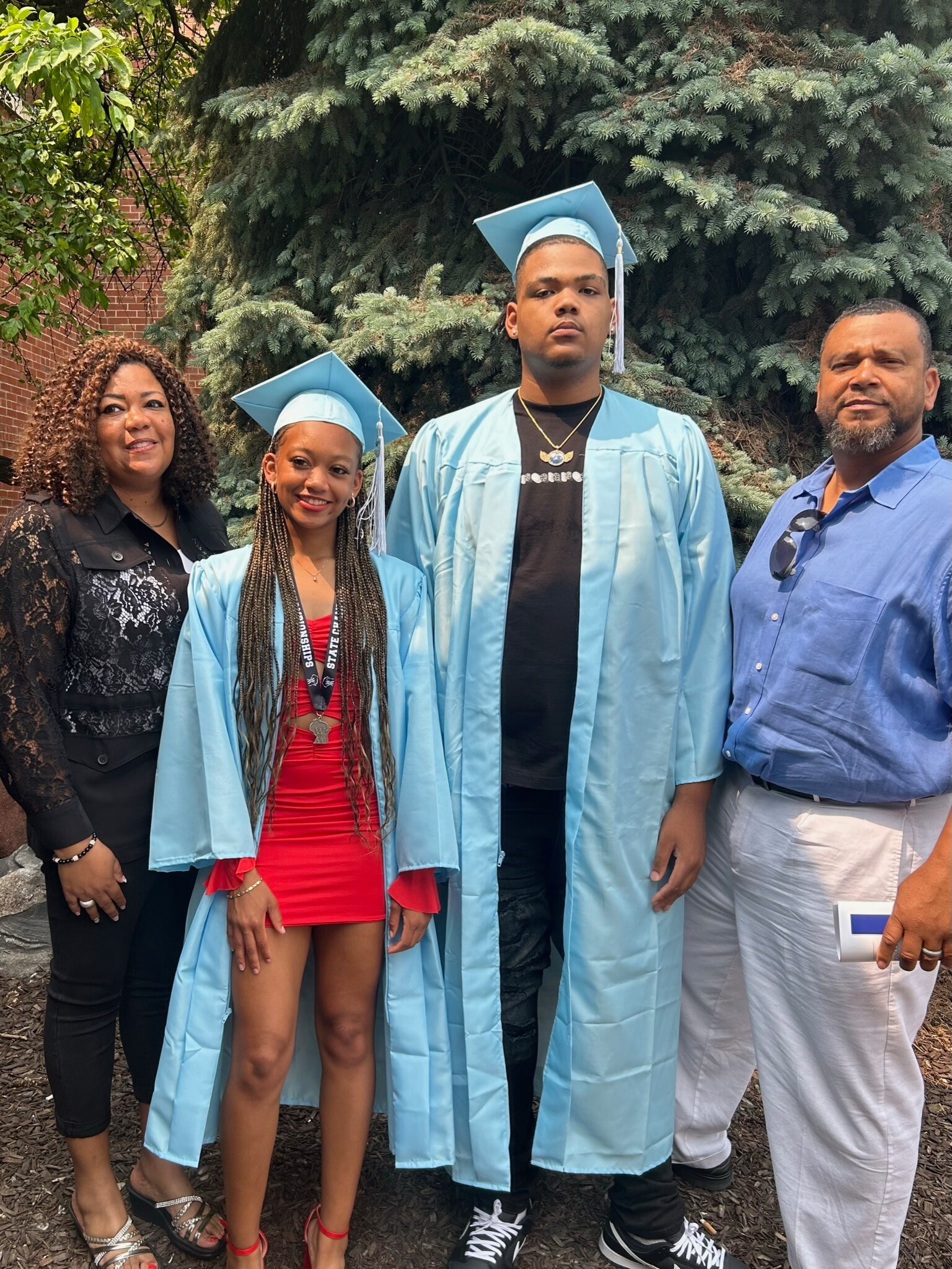 Four people pose outdoors after a graduation ceremony: two graduates in light blue gowns and caps, flanked by a woman in black and a man in a blue shirt.