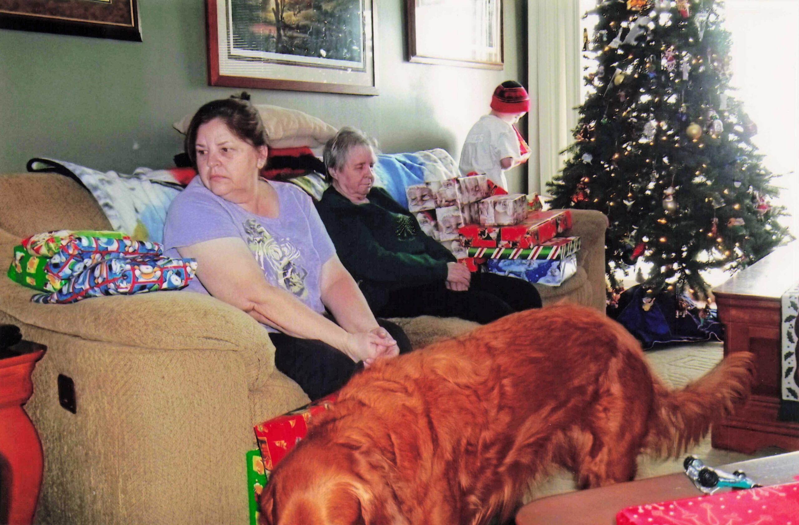Living room scene with two elderly women on a sofa amid wrapped Christmas gifts and a decorated tree nearby, including an orange dog in the foreground.