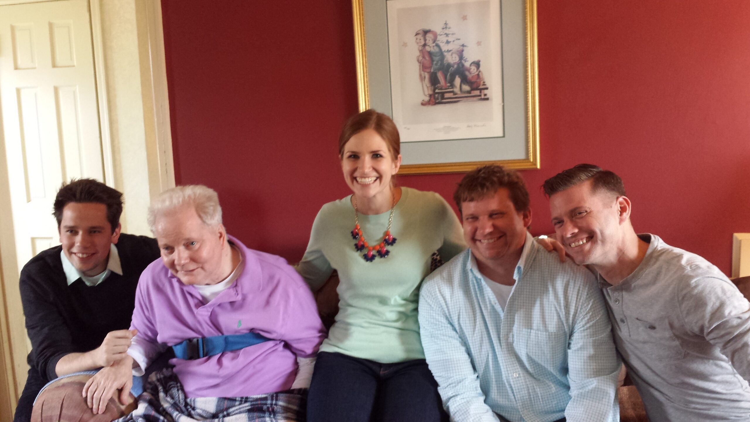 Five friends posing on a couch in a cozy living room with a red wall and framed art behind them, smiling at the camera.
