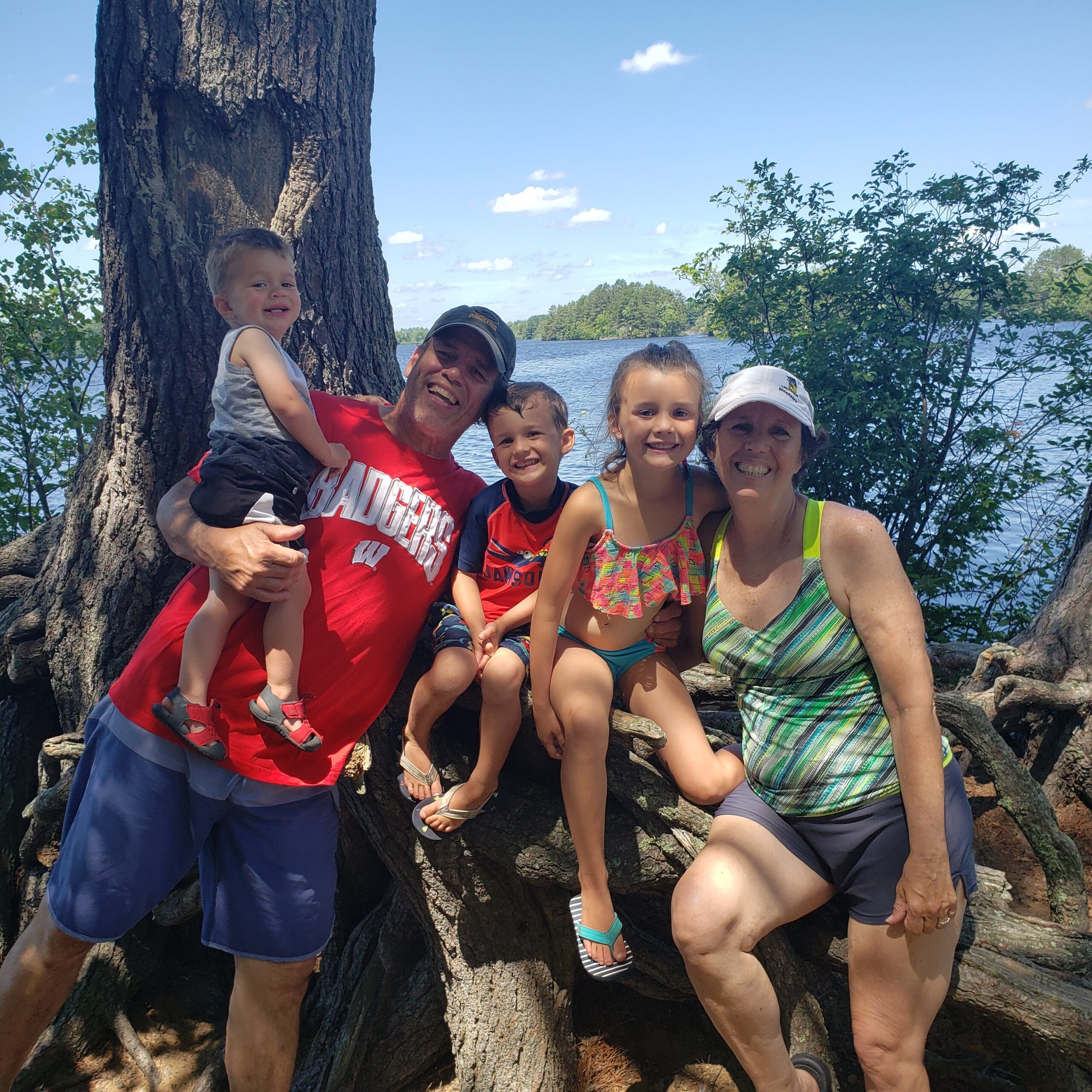 Family of six posing together by a large tree near a lake on a sunny day, smiling at the camera in casual clothing.