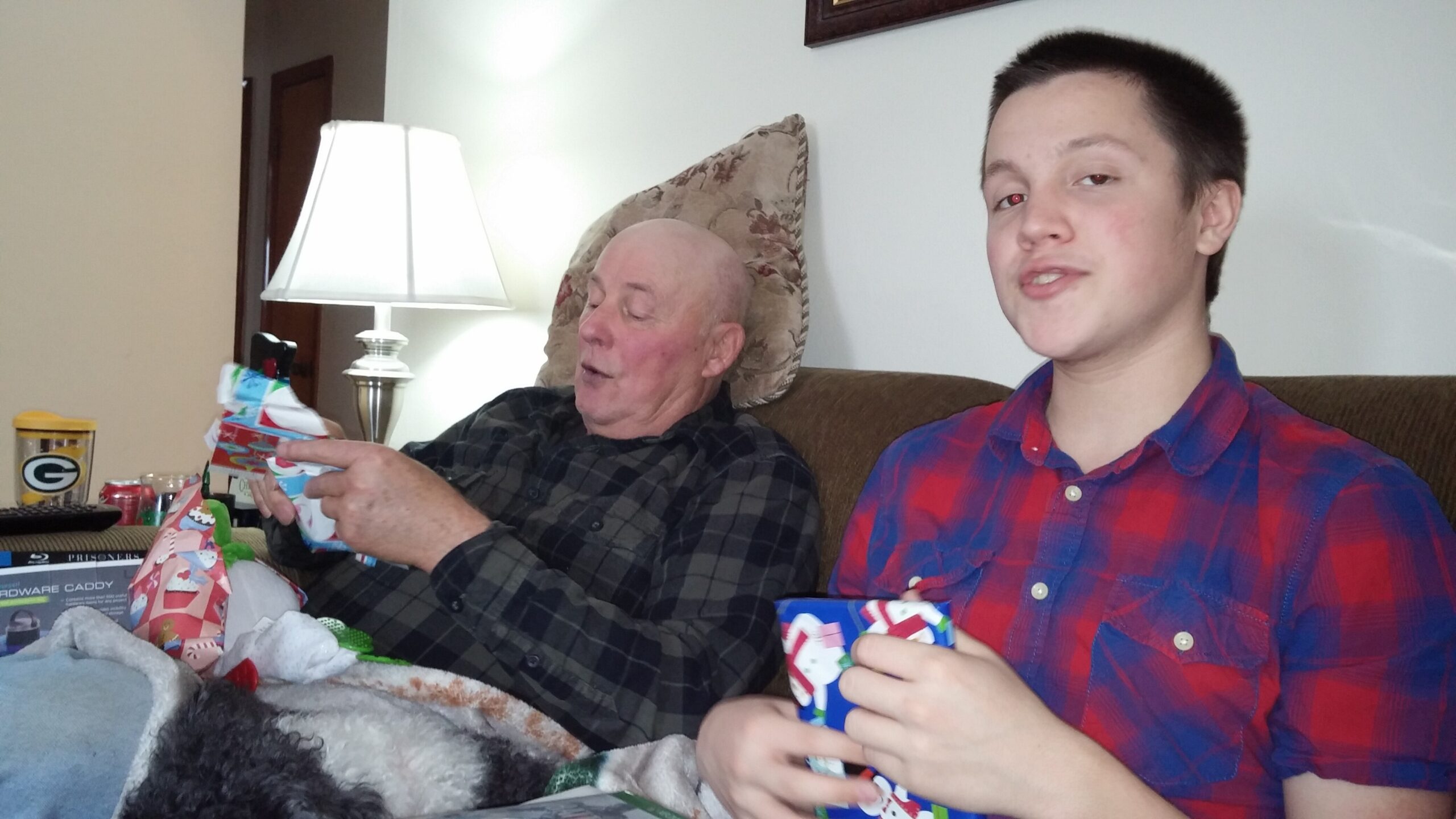 Two men sit on a couch unwrapping gifts, with wrapping paper scattered on a nearby table and a lamp in the background.