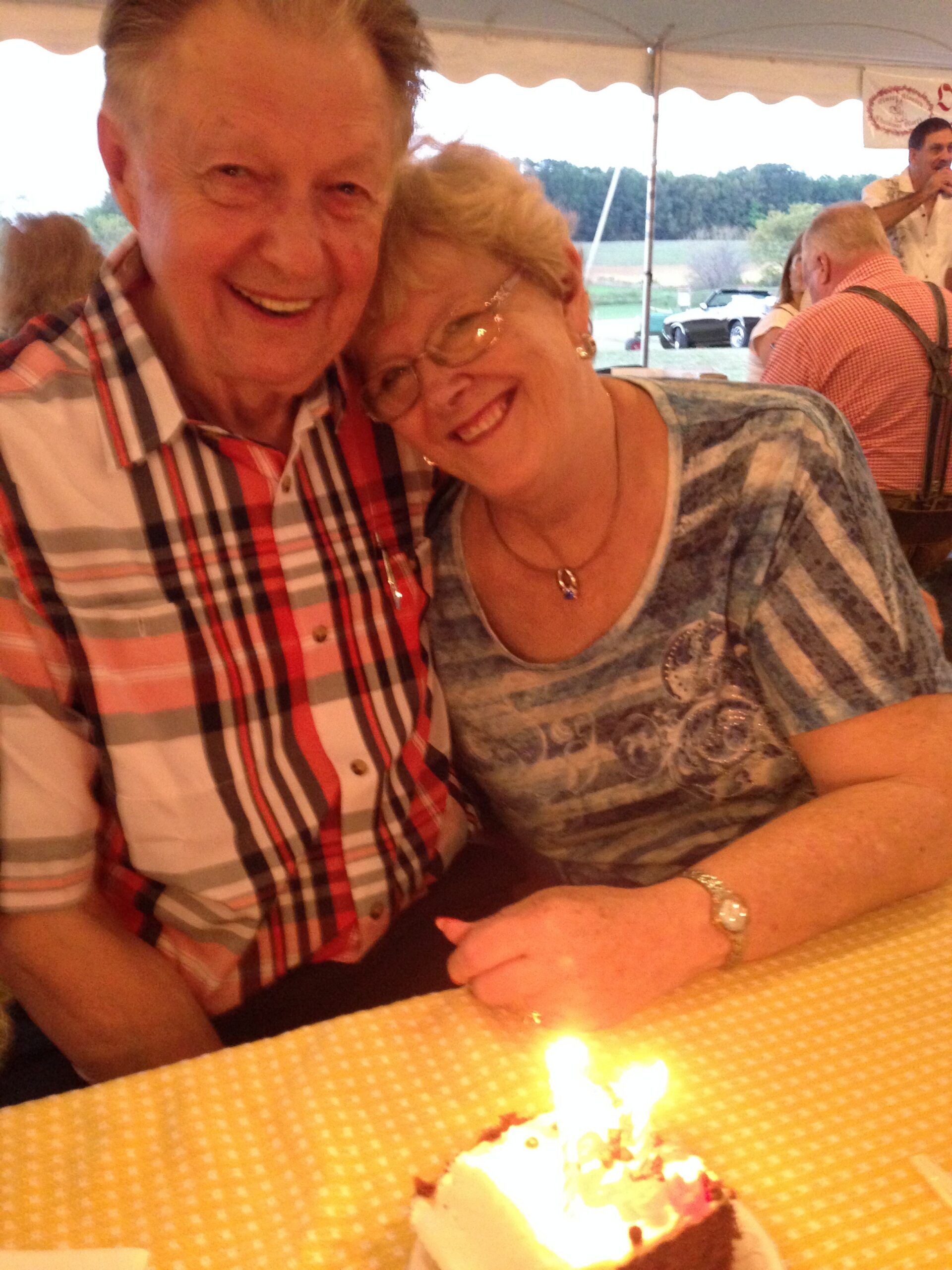 Senior man and woman smiling and leaning their heads together at a table with a lit birthday cake in front, outdoors under a tent.