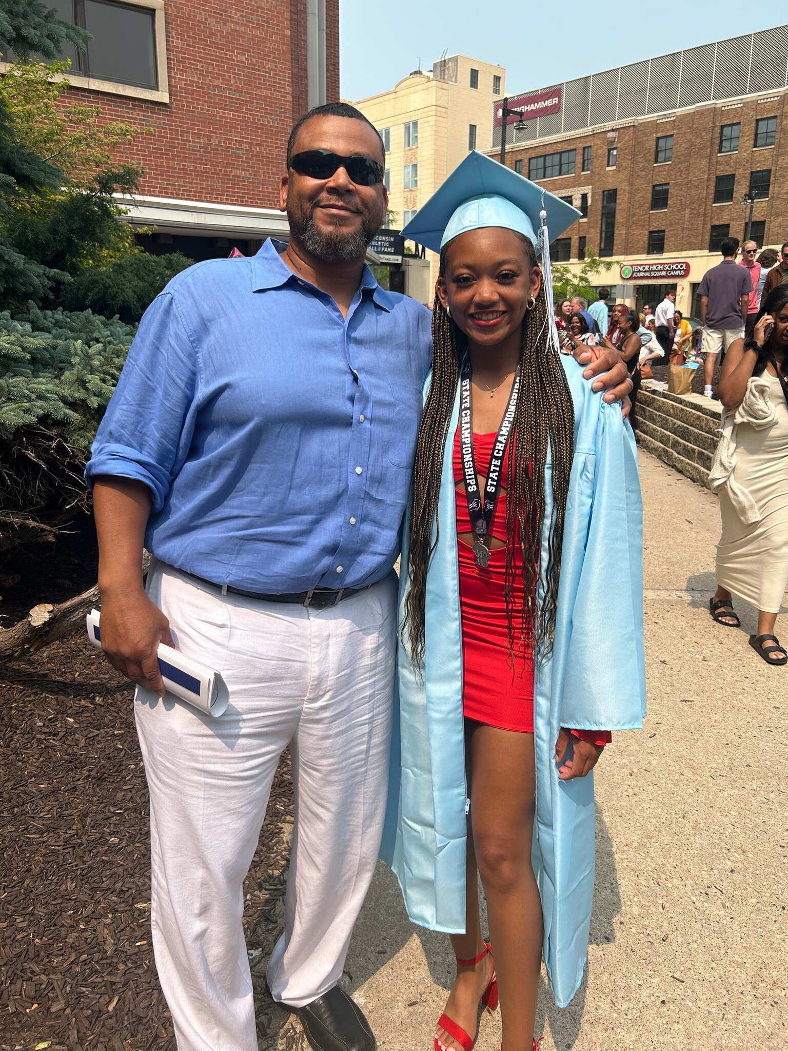 Man in a light blue shirt and white pants, wearing sunglasses, holds a rolled diploma, standing next to a graduate in a light blue cap and gown with a red dress and braided hair; both smile outdoors with a crowd in the background.