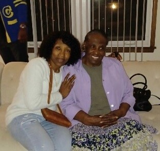 Two people sit on a white sofa, smiling for the camera: woman in a white cardigan and jeans with a brown purse, man in a lavender shirt and floral skirt.
