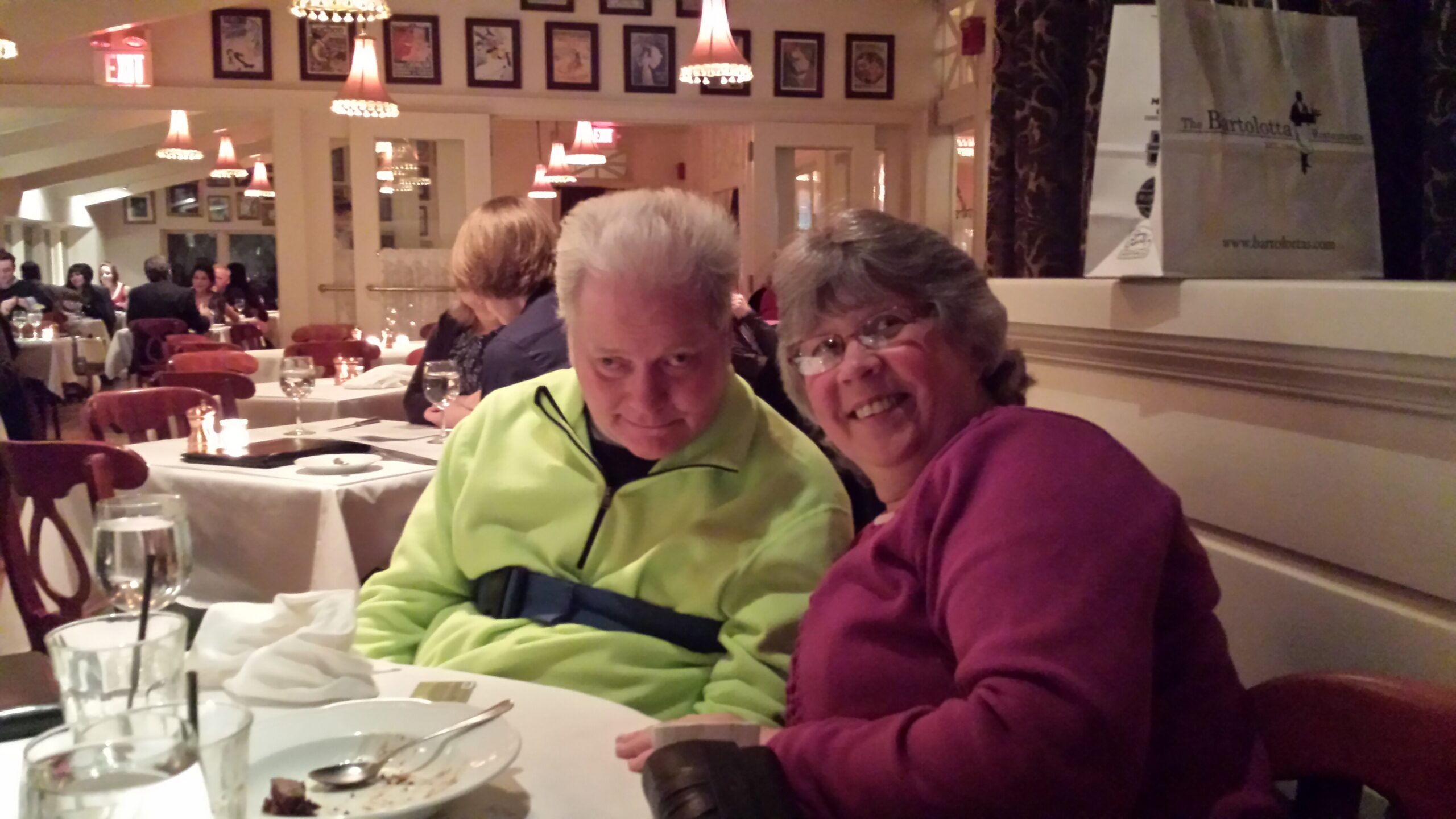 Smiling couple seated at a restaurant table in the foreground, with other diners and warm lighting in the background.