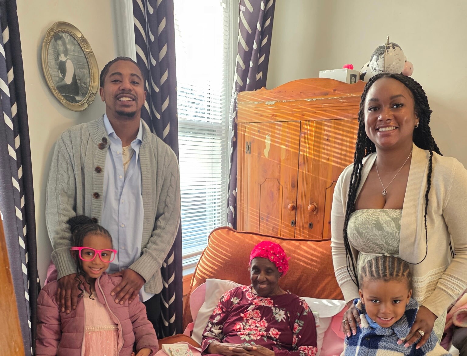 A smiling family posing in a living room: a man and a woman stand on either side of two seated women, with a child in pink glasses in the foreground.