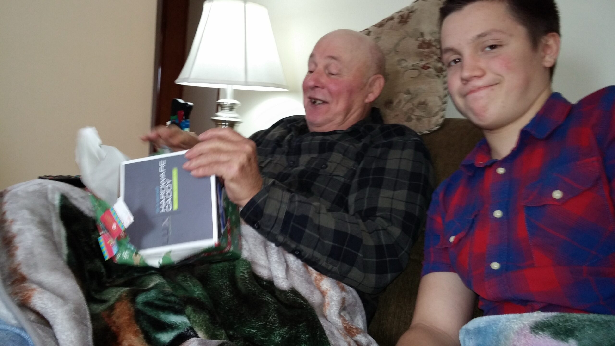 Two men sit on a couch as an older man opens a wrapped gift, smiling, while a younger man in a red plaid shirt sits beside him.