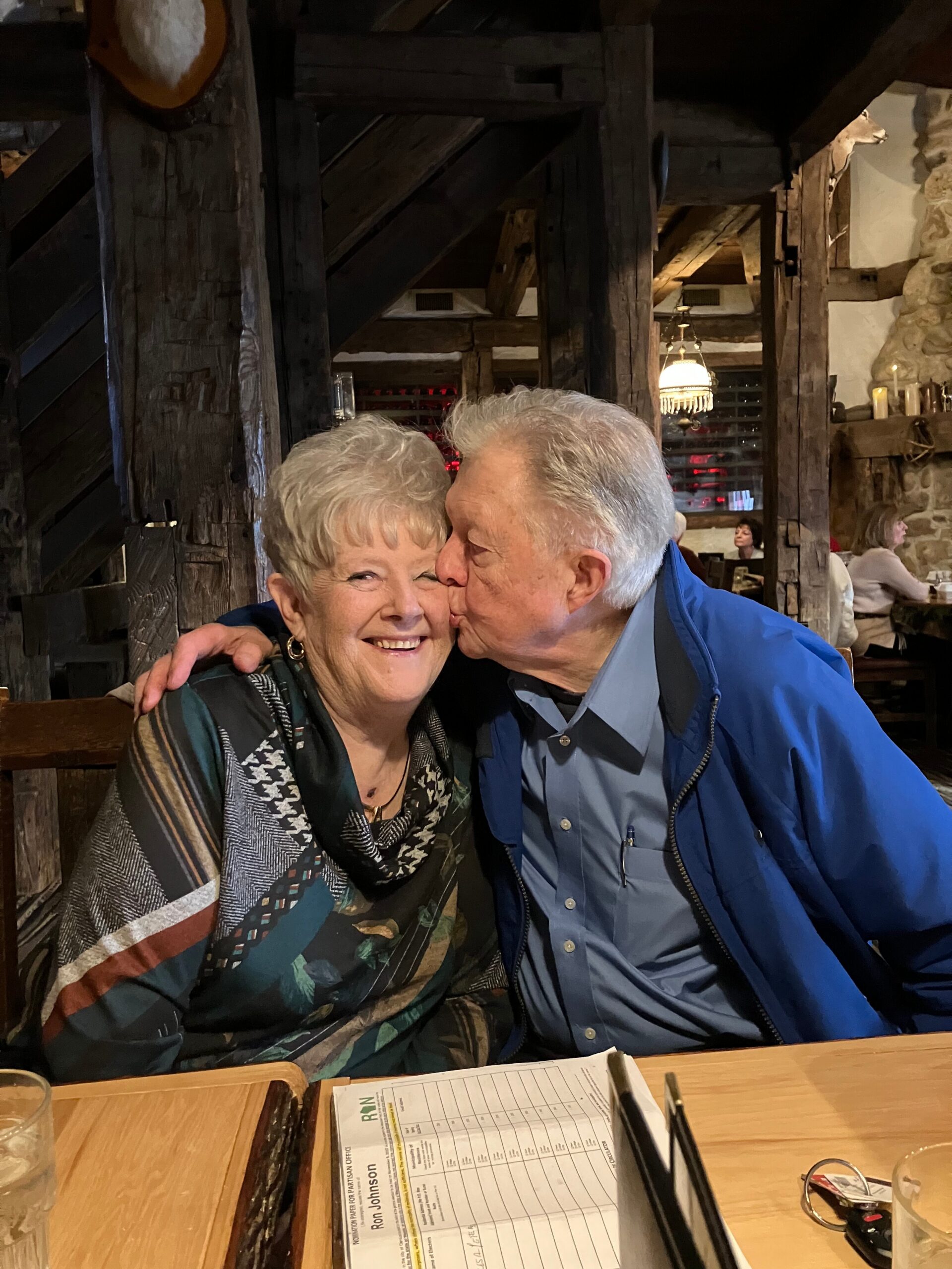 Elderly couple smiling as the man kisses the woman on the cheek at a rustic restaurant, wooden beams in the background.