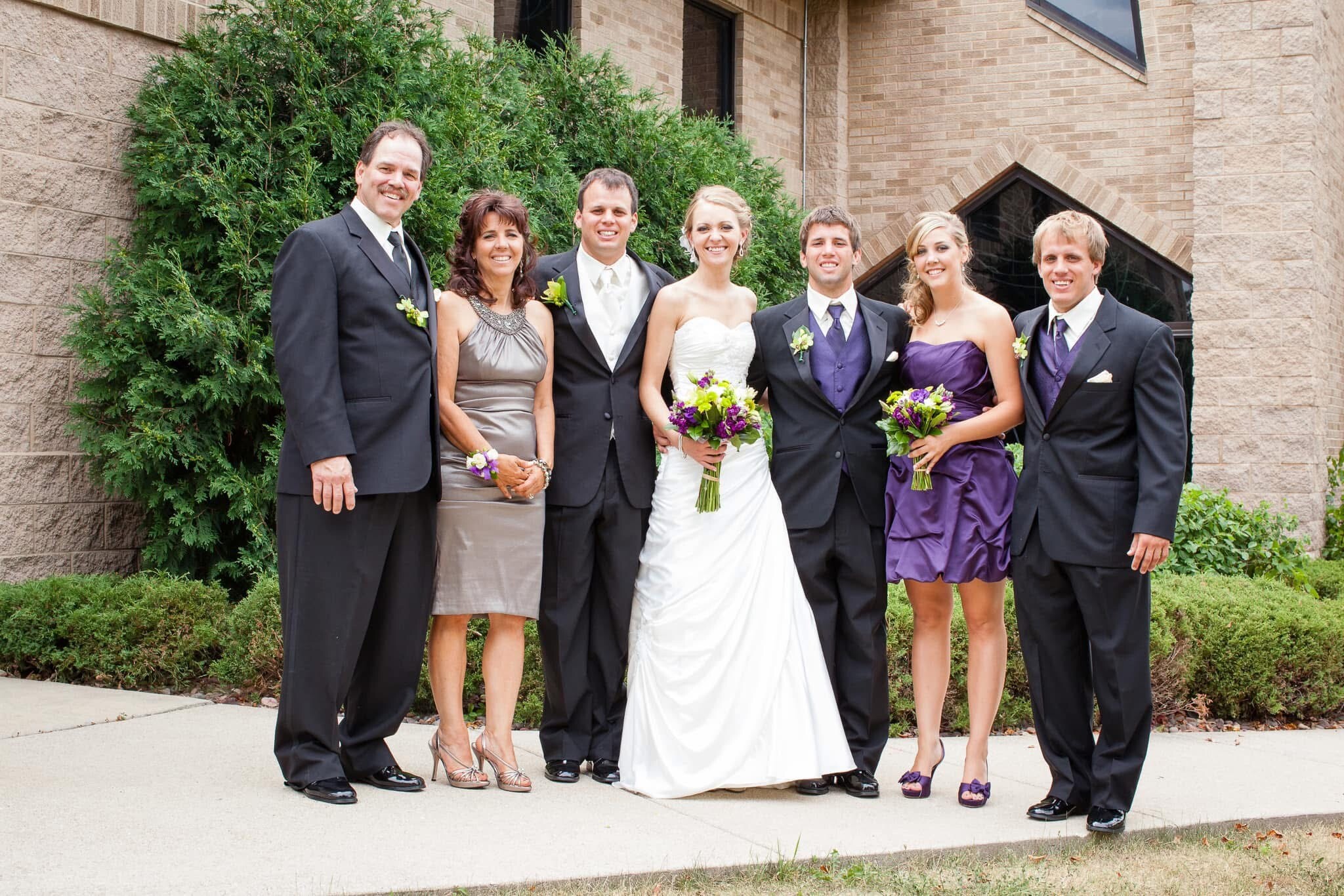 Wedding party photo: bride in white gown flanked by bridesmaids in purple dresses and groomsmen in black suits, outside a brick building with greenery to the left.