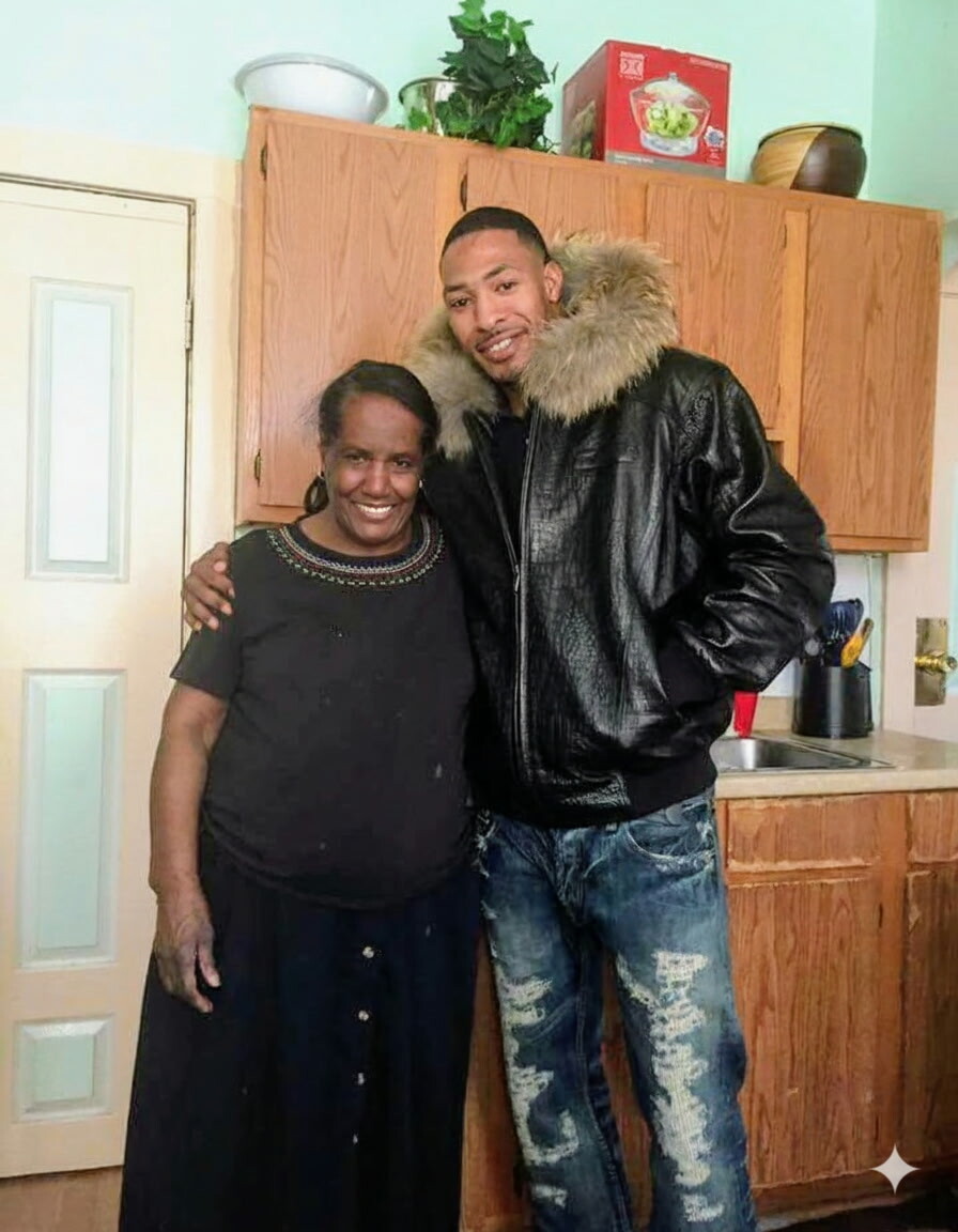 Two people pose together in a home kitchen, smiling; woman wears a dark top, man wears a black fur-lined jacket.