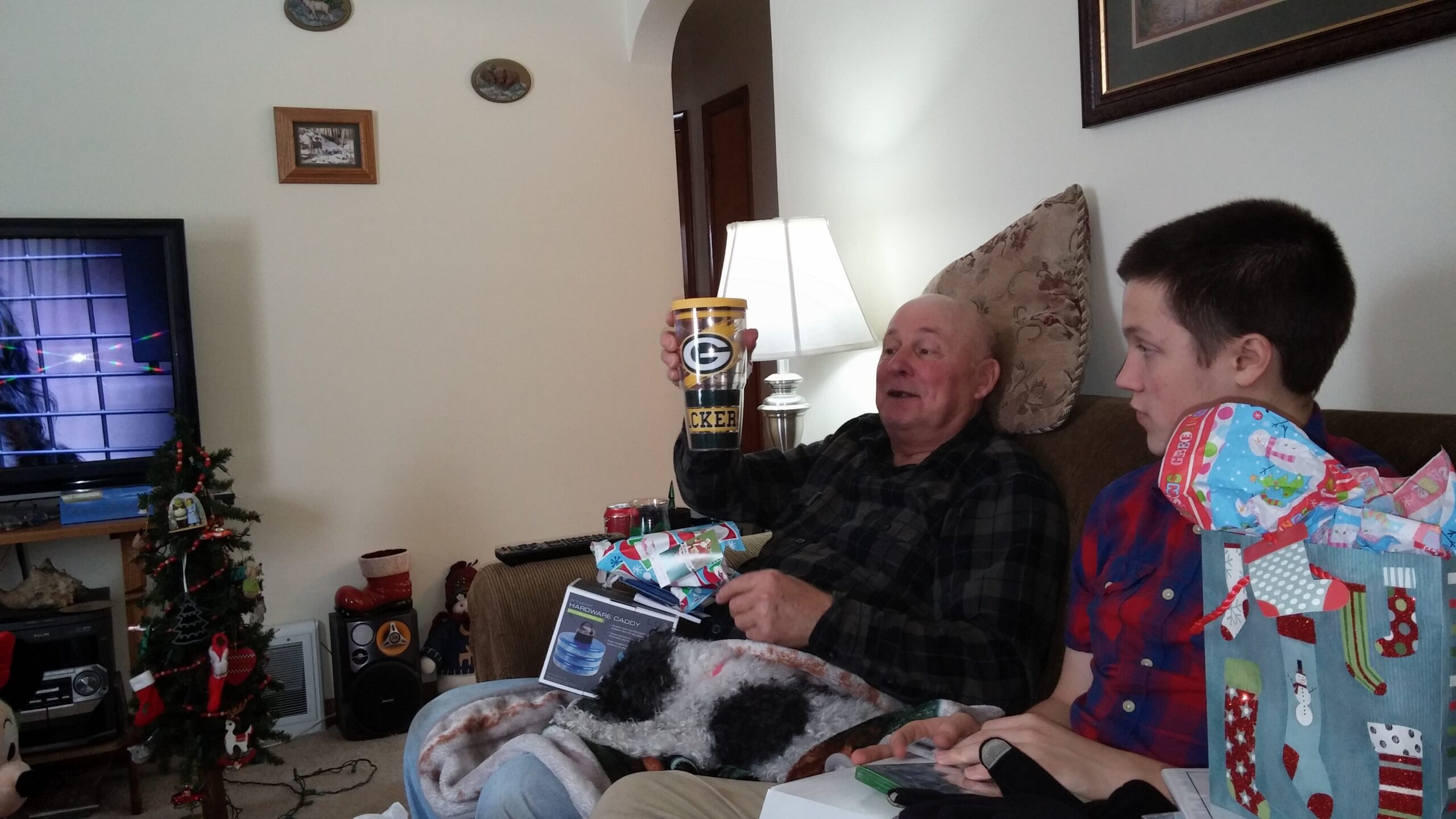 Older man on a sofa raises a Packers tumbler as a younger man sits beside him among wrapped gifts and a Christmas tree in a living room.