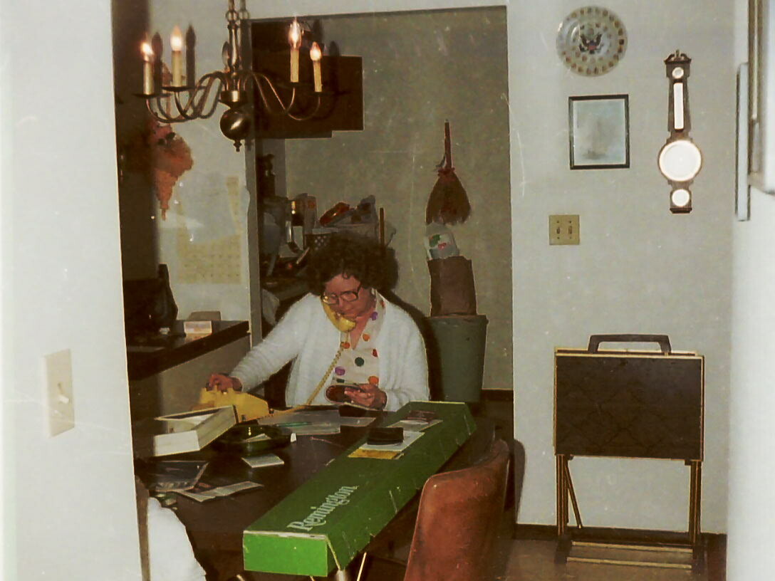 Woman with glasses sits at a cluttered desk indoors, talking on a yellow landline phone and writing notes.